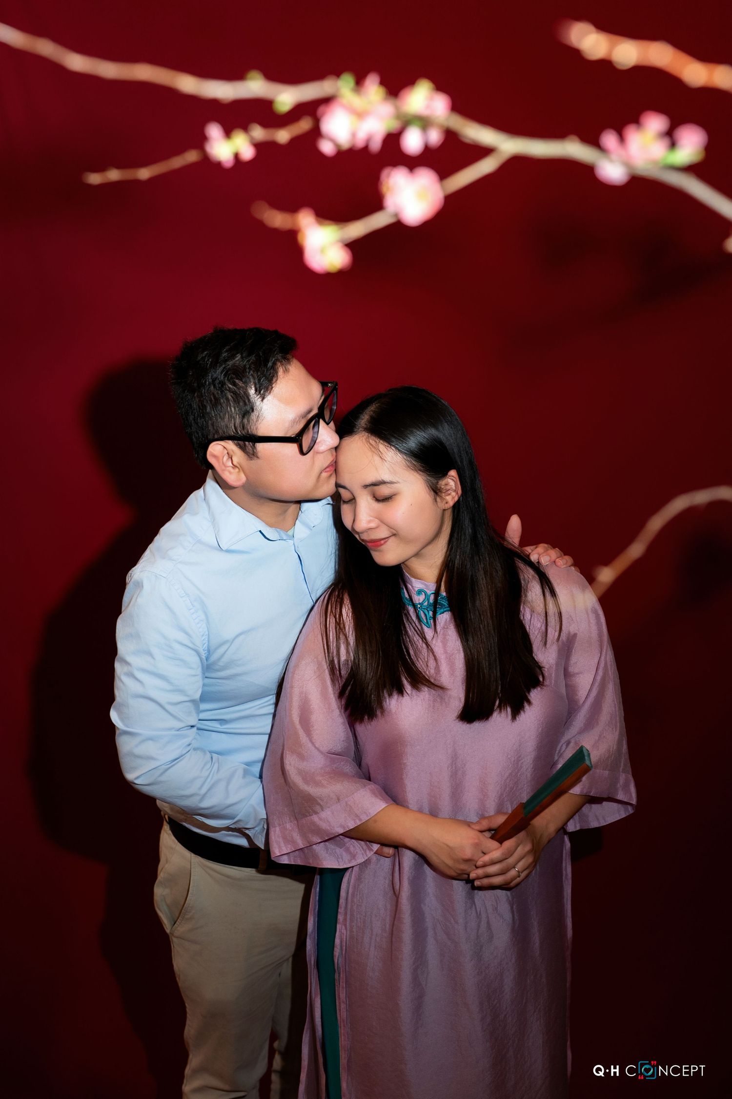 Couple embraces in front of red backdrop with delicate cherry blossom branches.