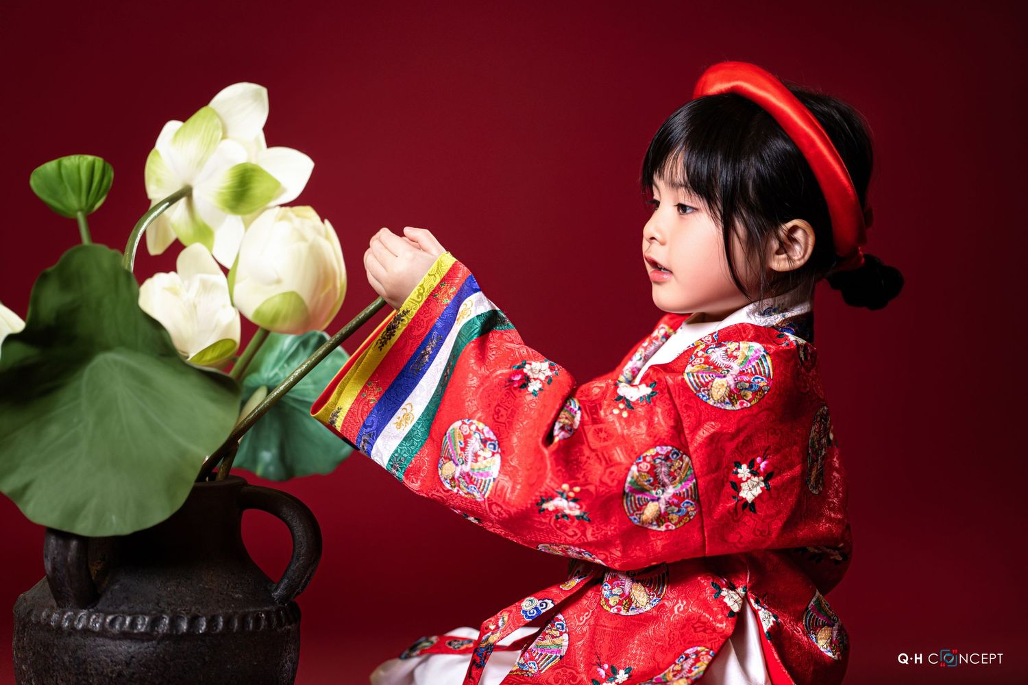 A child in traditional red Asian dress poses with lotus flowers against a deep red backdrop.