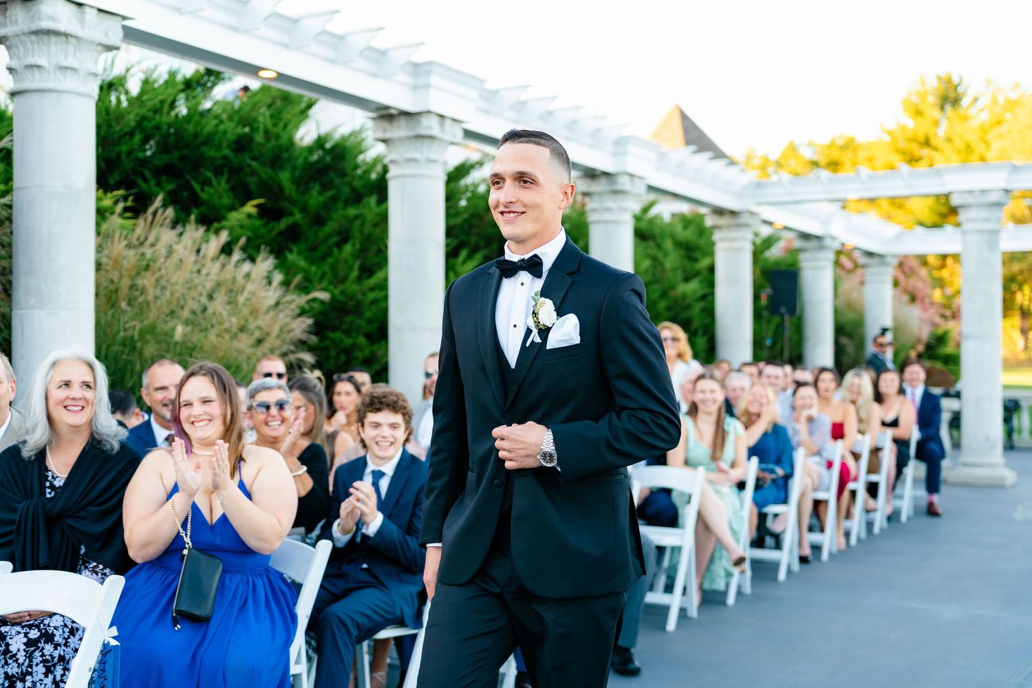 Wedding ceremony guests seated in white chairs under elegant white pergola columns during outdoor celebration.