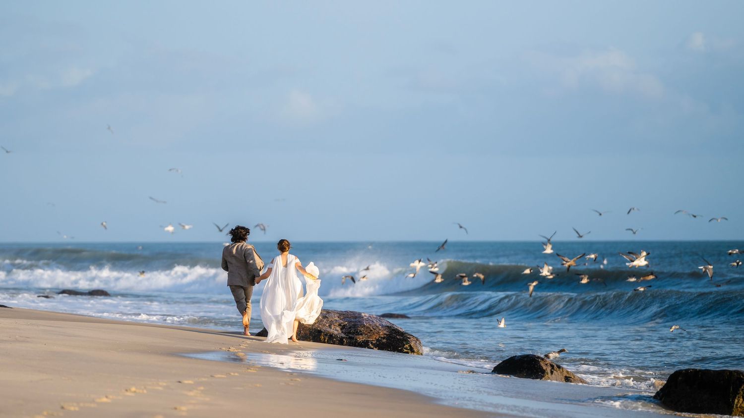 Panoramic view of a sandy beach with waves crashing on the shore and seagulls flying over the ocean on a sunny day.