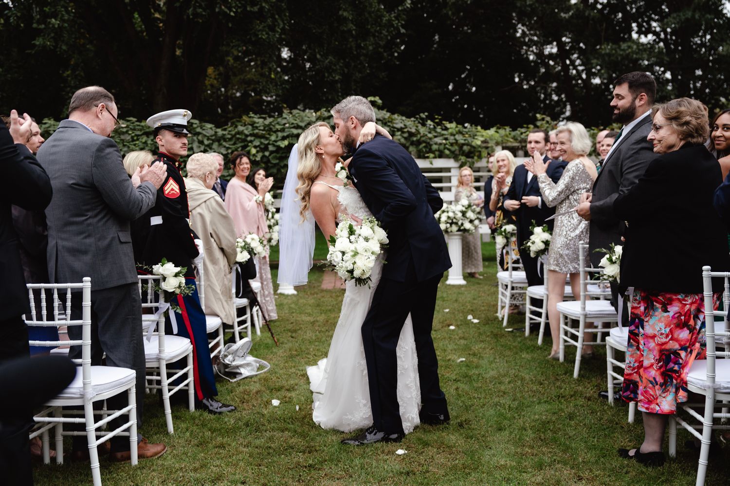 Wedding guests stand in rows on a grassy lawn during an outdoor ceremony with white chairs arranged in aisles.