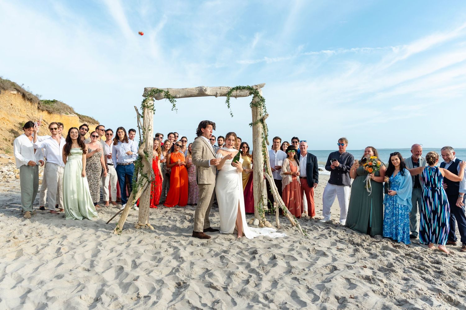 Large group of wedding guests gathered on a beach with dramatic cliffs and blue sky overhead.
