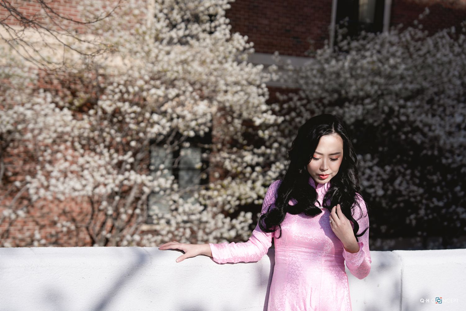 Woman in pink dress poses near blooming white cherry blossom trees in spring sunlight.