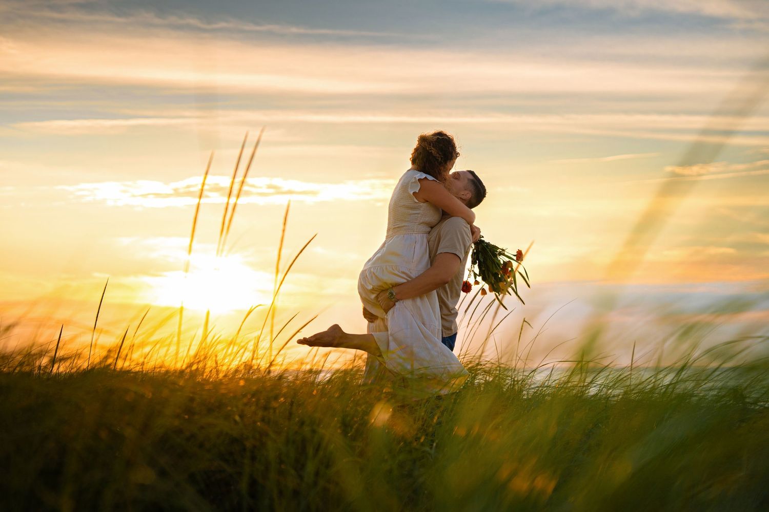 Romantic couple embraces in golden field at sunset with sunbeams streaming through tall grass.