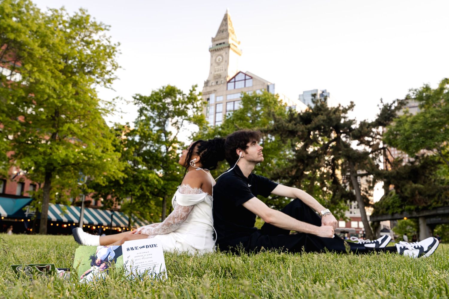 Couple sitting back-to-back on grass in Boston park with historic clock tower backdrop on a summer day.