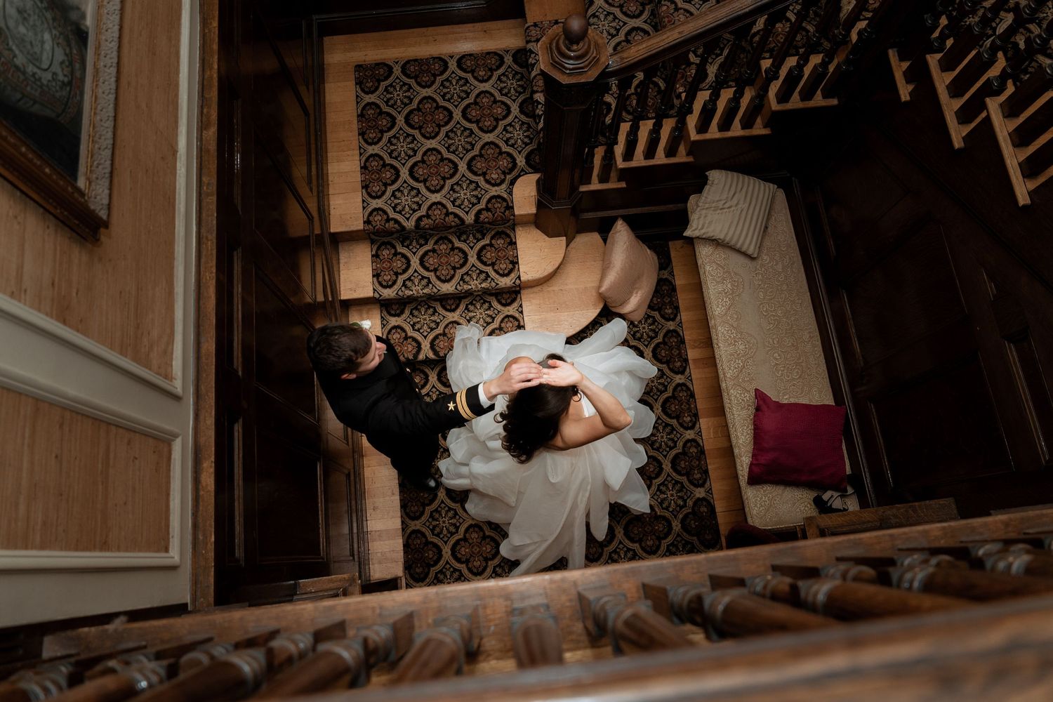 A couple in wedding attire lies on ornate vintage stairs in a romantic, moody atmosphere captured from above.