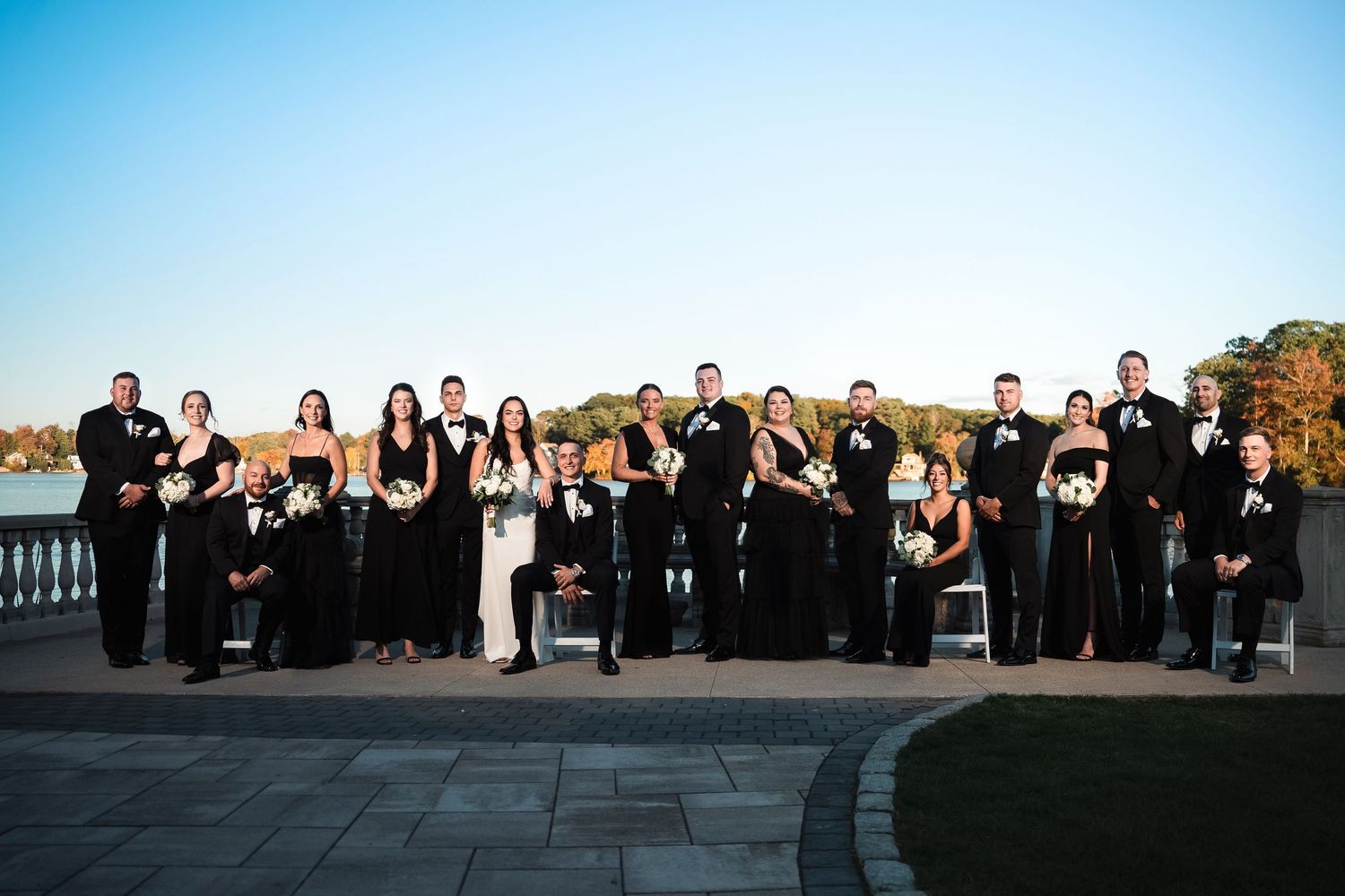 A wedding party dressed in formal black attire poses together on a stone patio overlooking a lake during autumn.