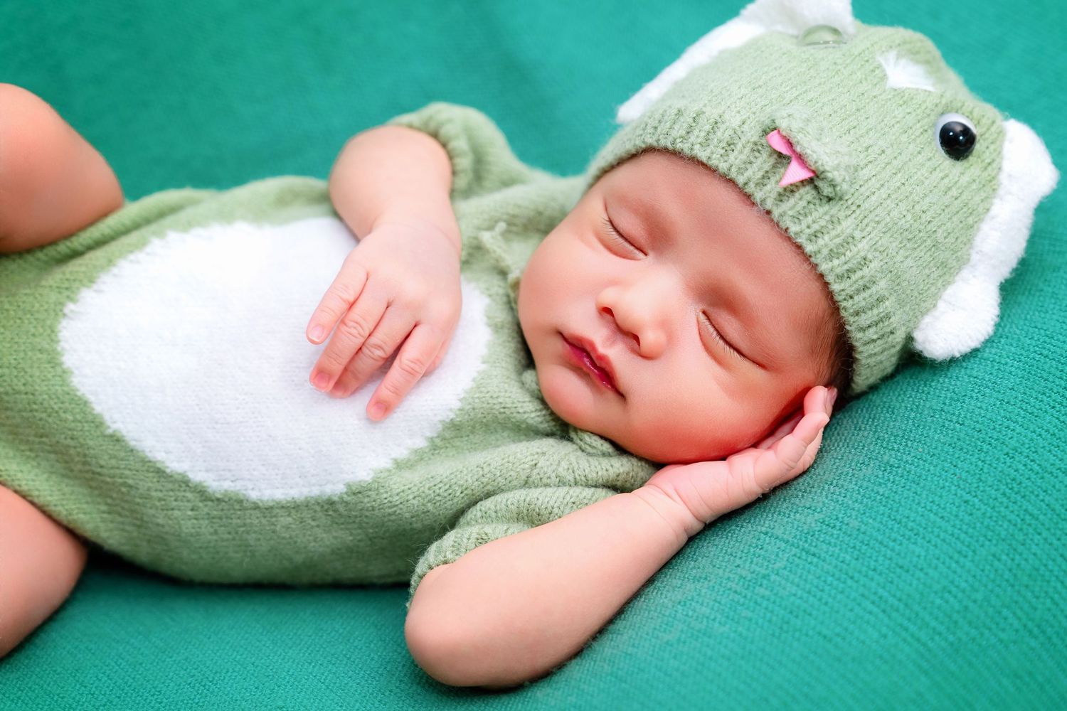 A sleeping newborn in mint green knit outfit with bunny ear hat on turquoise backdrop.