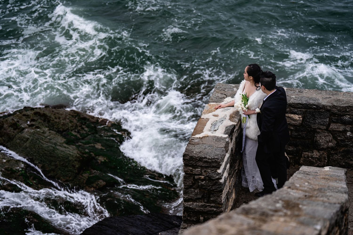 Person in black sits on rocky cliff edge overlooking turbulent ocean waves crashing against the coastline.