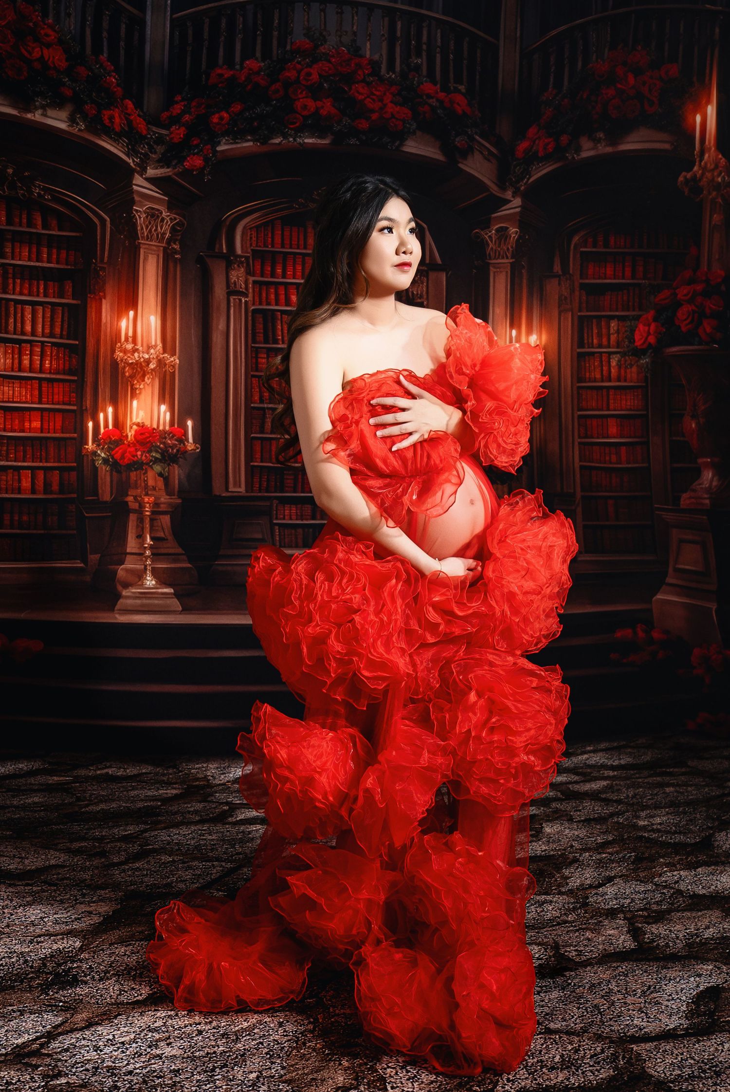 Dramatic maternity photos in library setting featuring flowing red tulle dress against dark bookshelves.