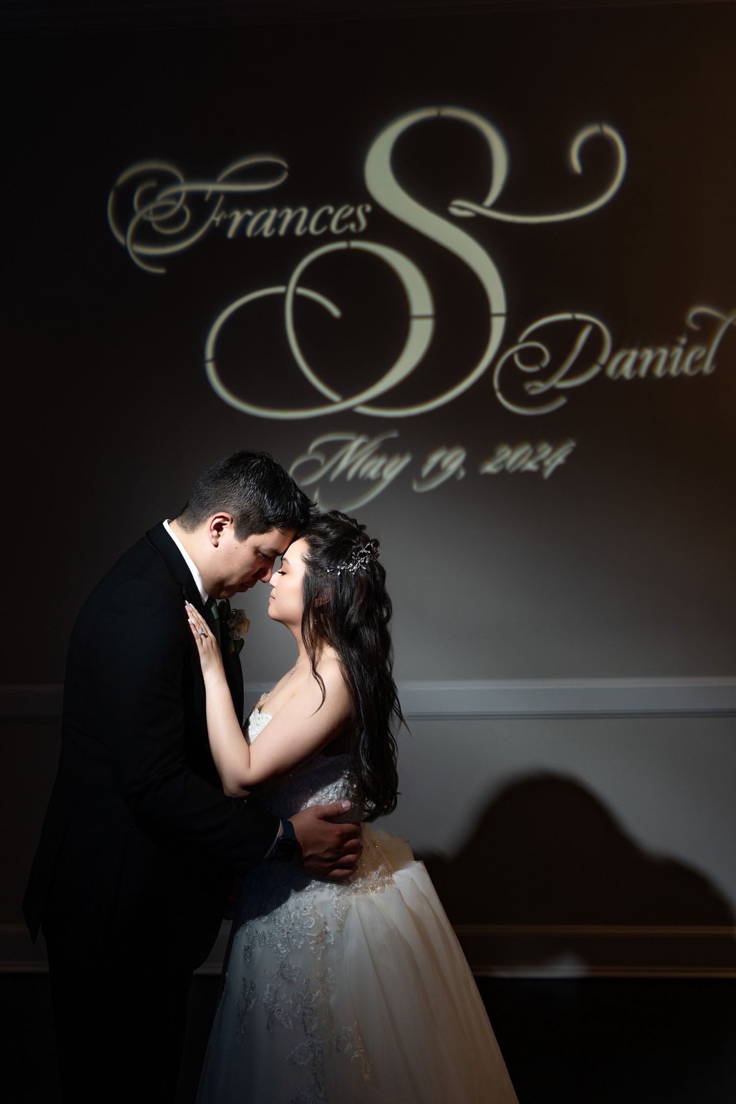 Dramatic silhouette of newlyweds kissing with elegant script monogram projection in background.