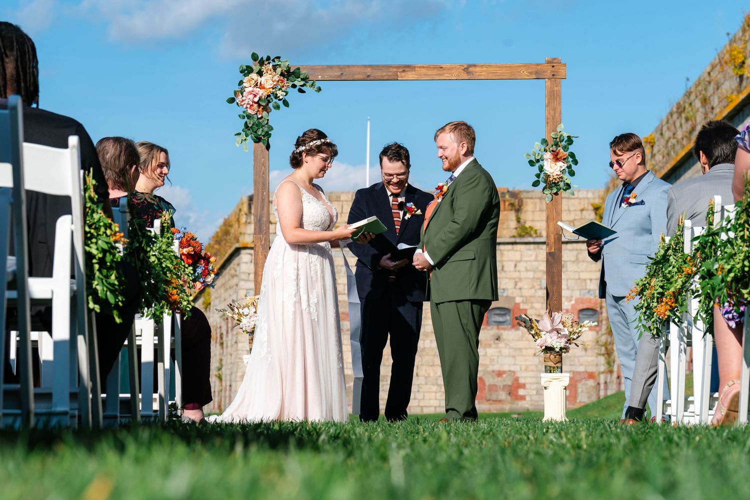 Outdoor wedding ceremony under wooden arch decorated with floral arrangements and greenery.