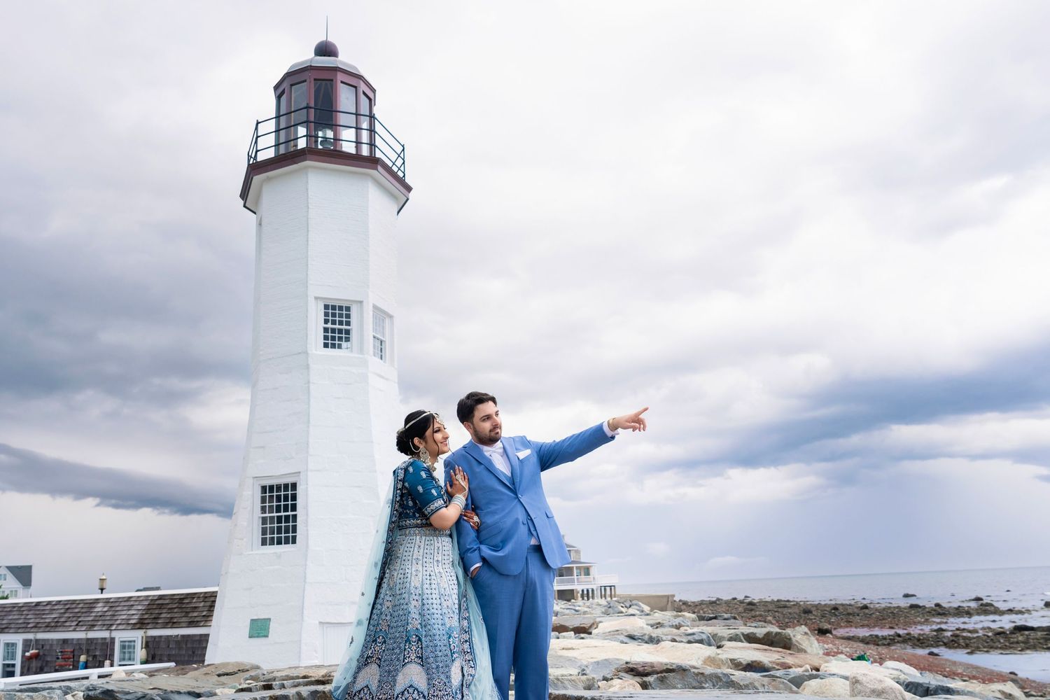 Couple in wedding attire pointing at view from white lighthouse under cloudy sky.