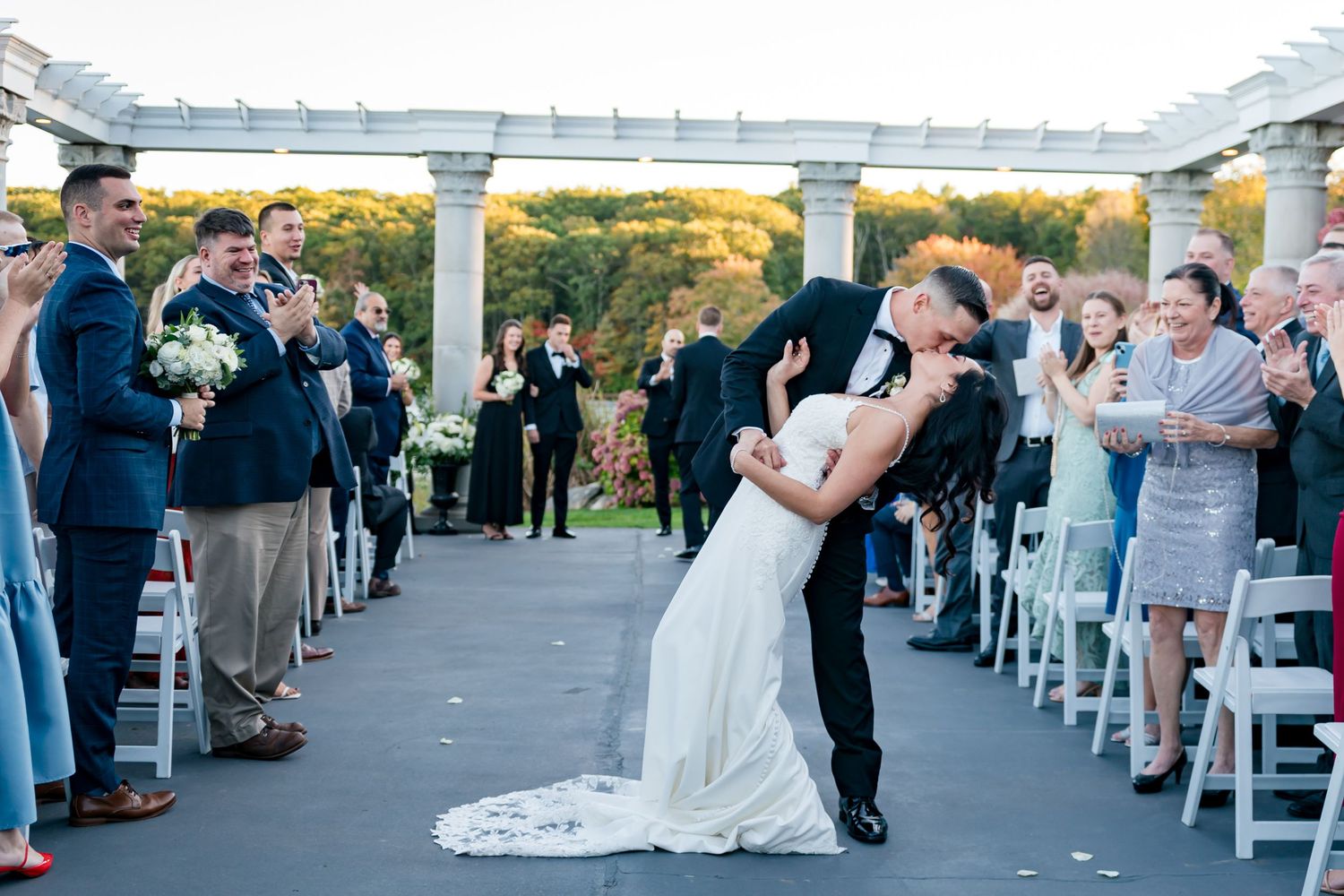 Outdoor wedding ceremony under white pergola with guests seated on white chairs.