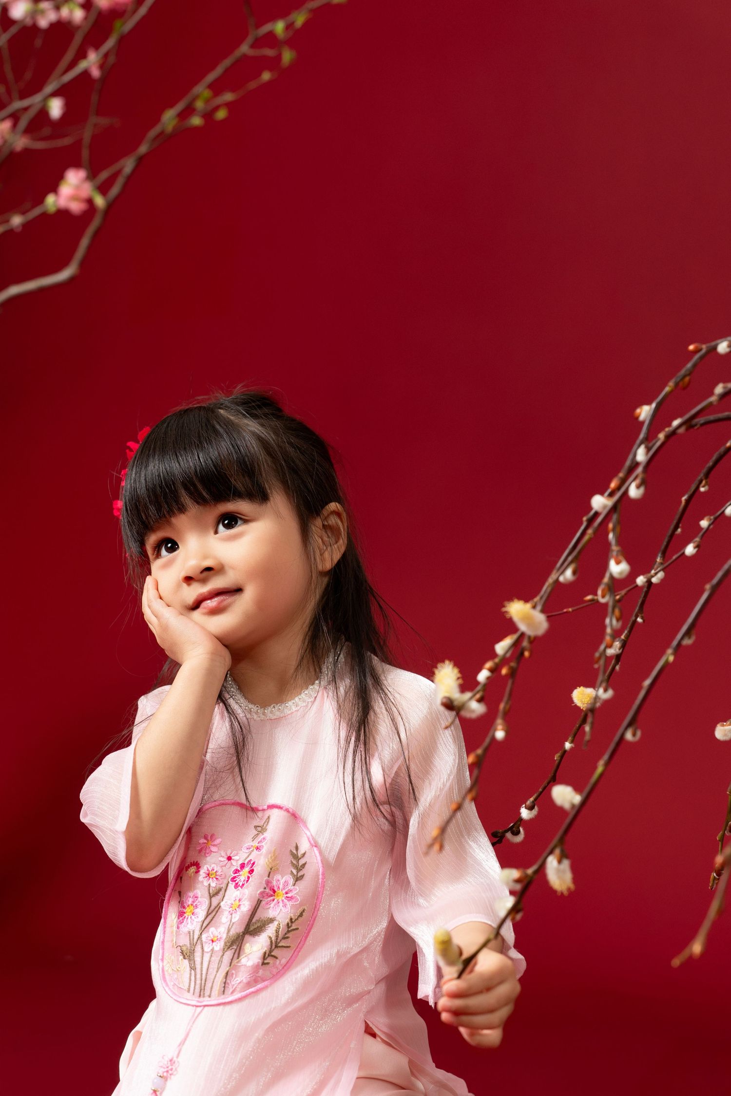 A child in a pink dress poses playfully against a deep red background with delicate cherry blossoms in view.