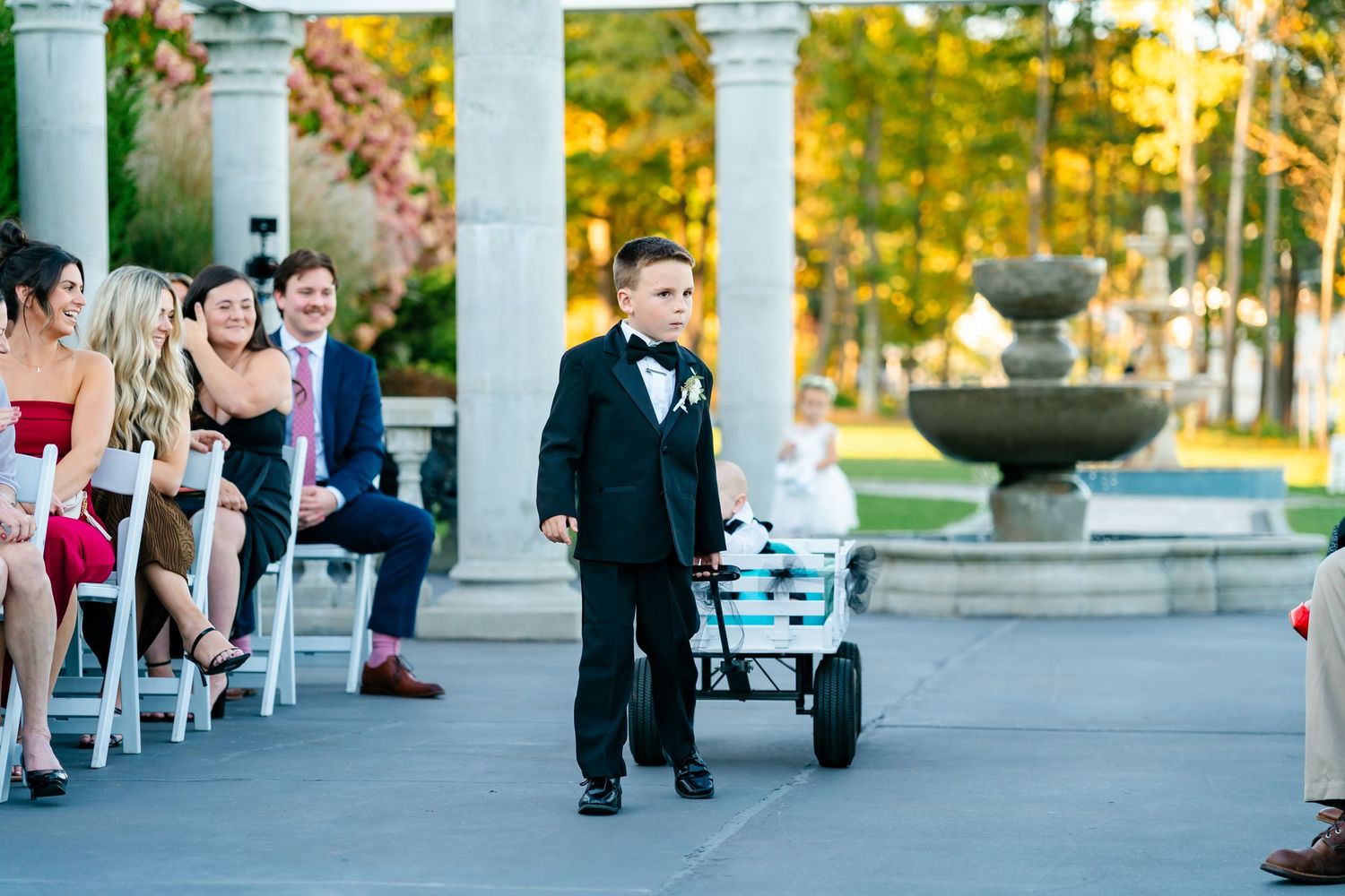 Young ring bearer pulls wagon down aisle during outdoor wedding ceremony.