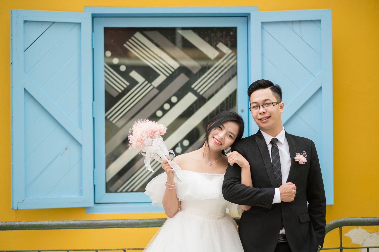 A wedding couple poses against a vibrant yellow wall with blue window shutters in an artistic composition.