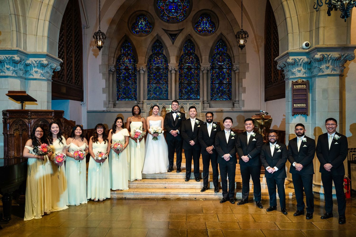 Wedding party lined up in formal attire inside historic church with stained glass windows.