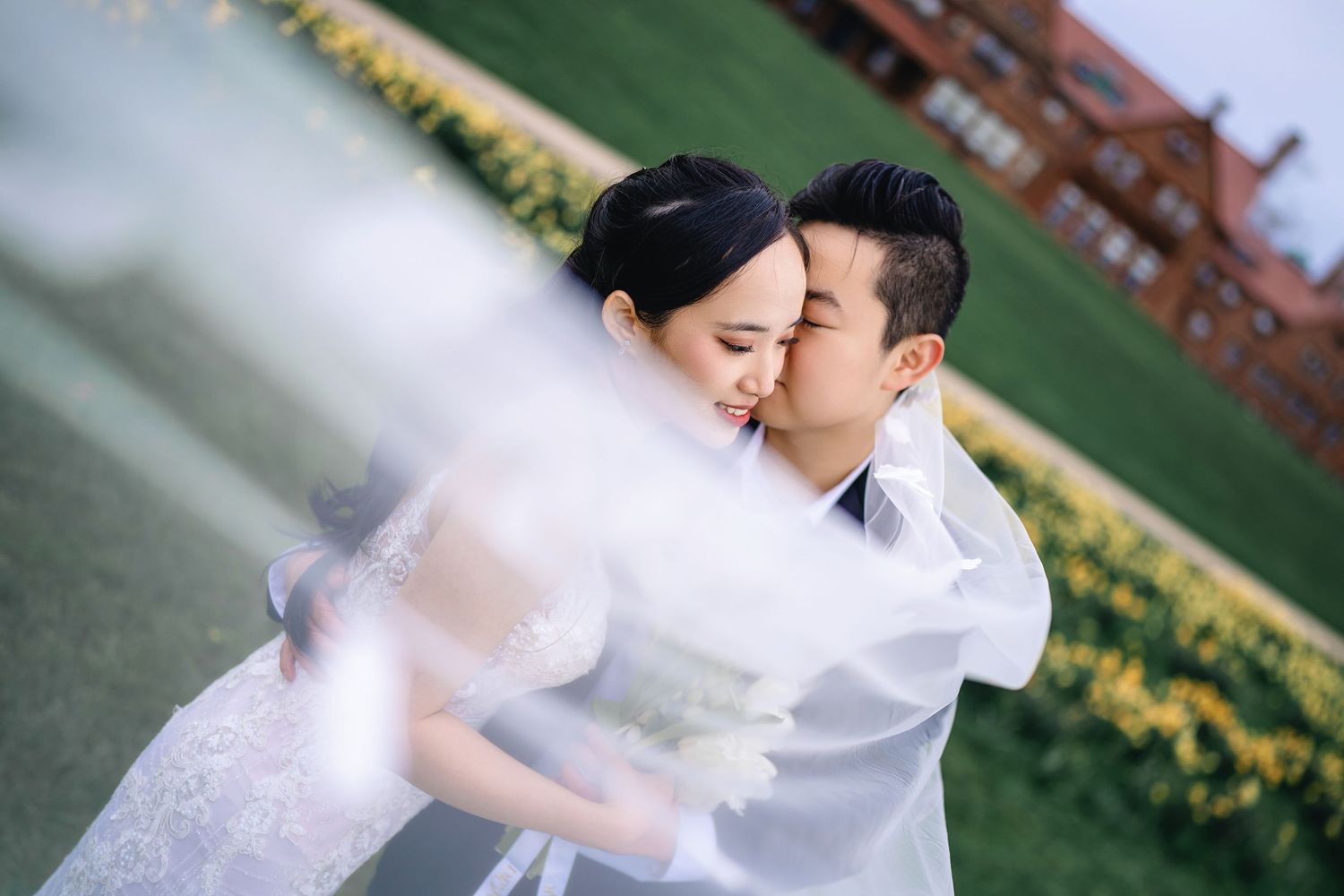 A couple in wedding attire embraces romantically in a garden setting with yellow flowers in the background.