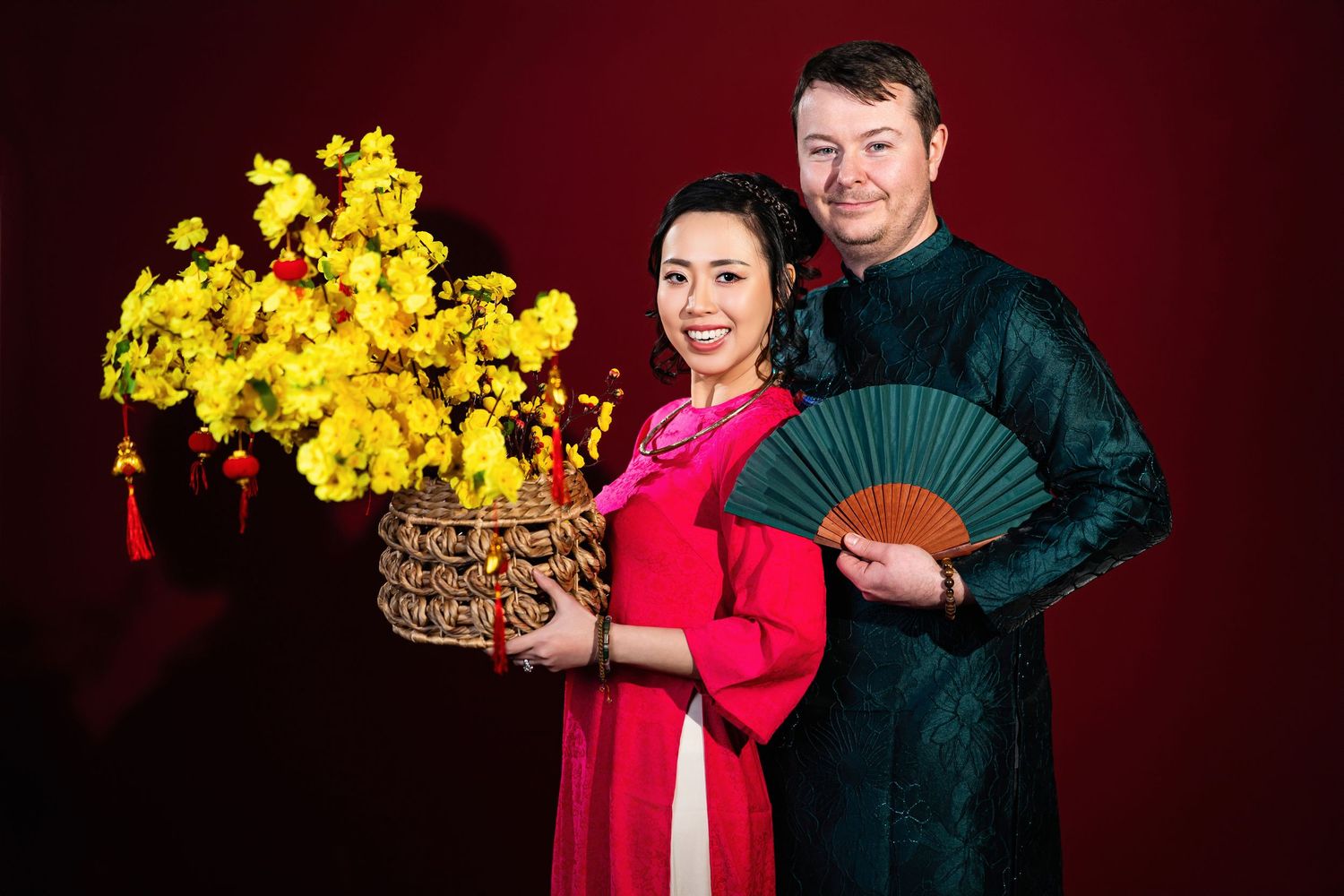 Couple in red and black outfits pose playfully with yellow chrysanthemums against burgundy background.