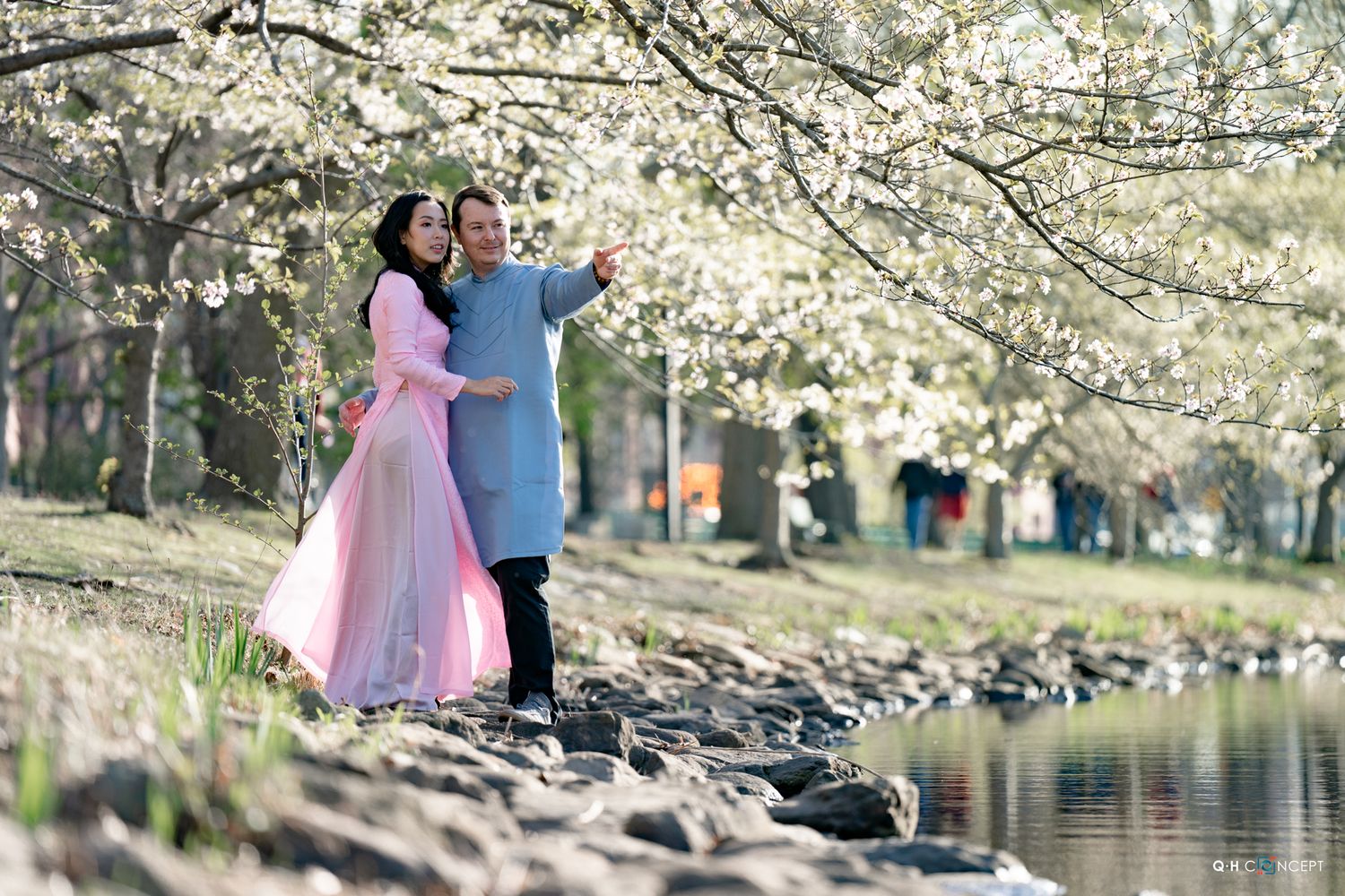 Couple in pink and blue outfits posing under blooming white cherry blossom trees by a serene river.