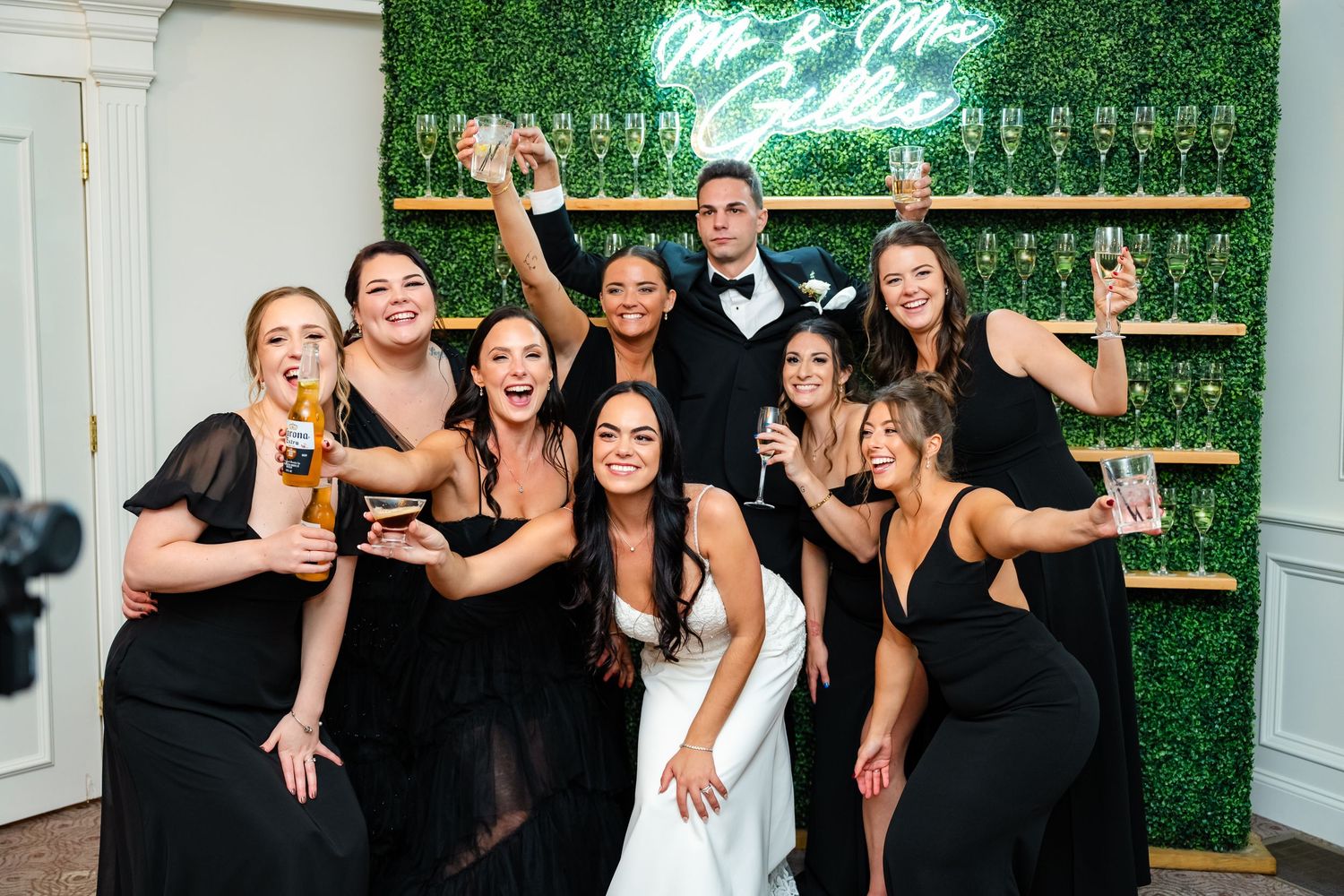 Bridal party in black dresses poses playfully with drinks against a green wall display.