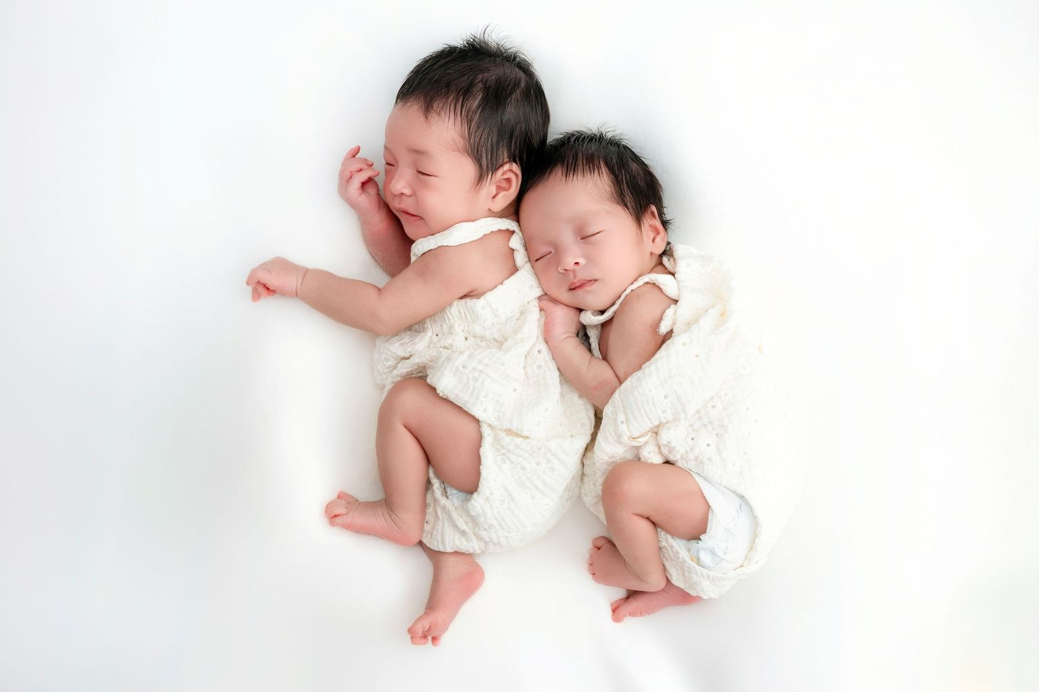 Two sleeping newborn twins wearing white outfits on white background.