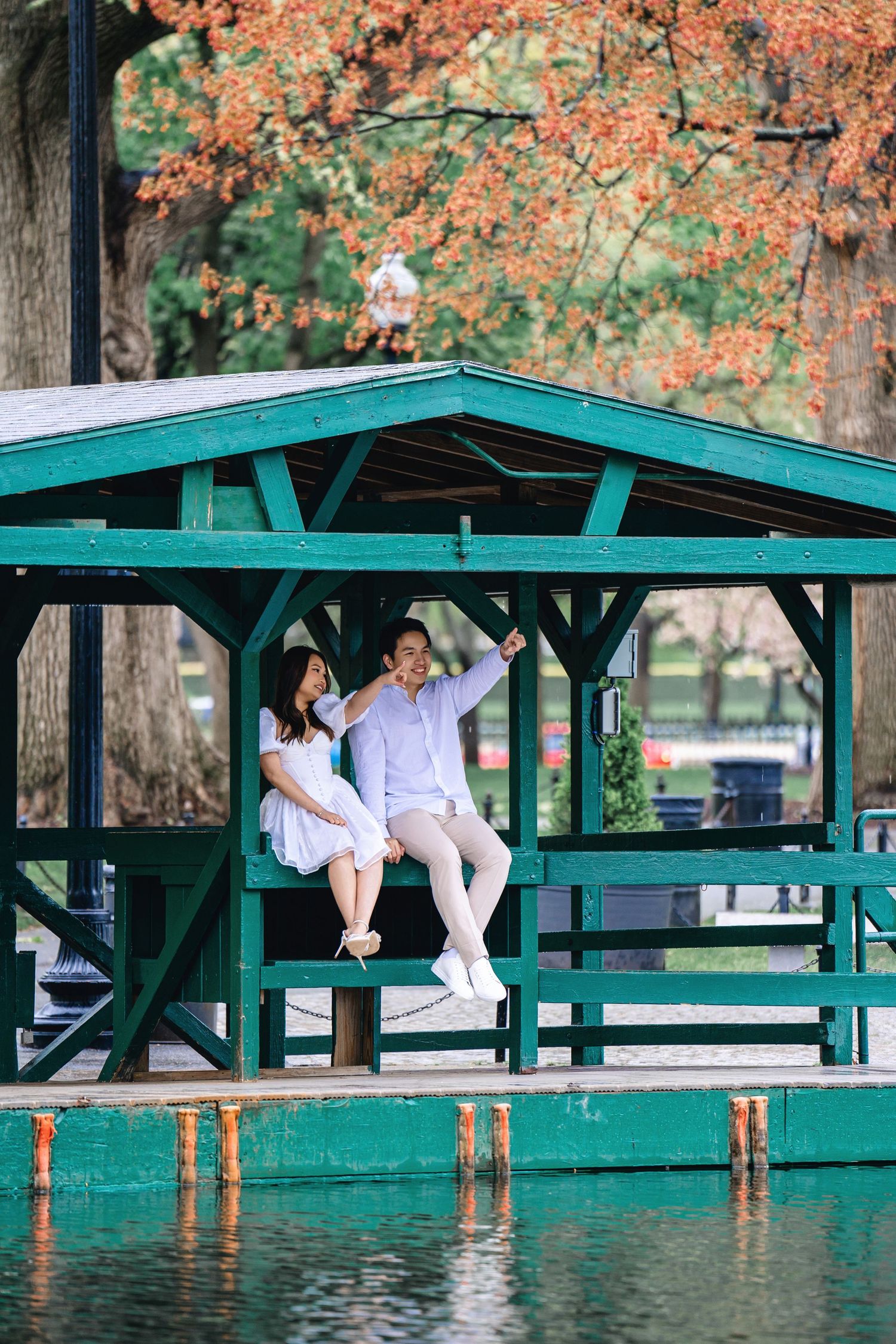 A couple in white outfits shares a romantic moment on a green wooden pier surrounded by autumn foliage.