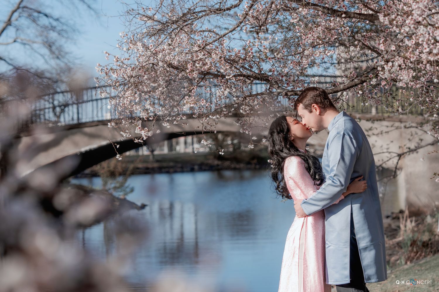 Couple embracing under blooming cherry blossom trees beside a tranquil pond during springtime.