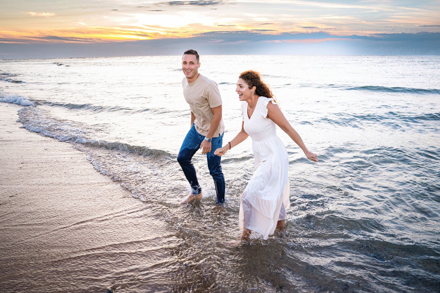 Joyful couple runs through ocean waves at beach during colorful sunset.