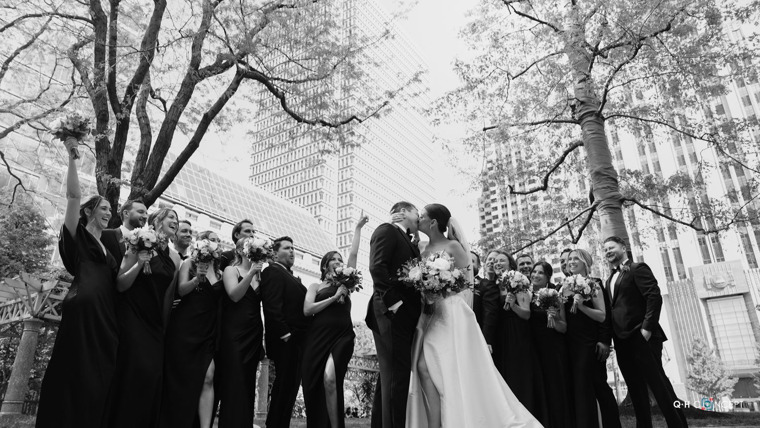 A black and white wedding photo showing a couple surrounded by their wedding party in an outdoor urban setting.