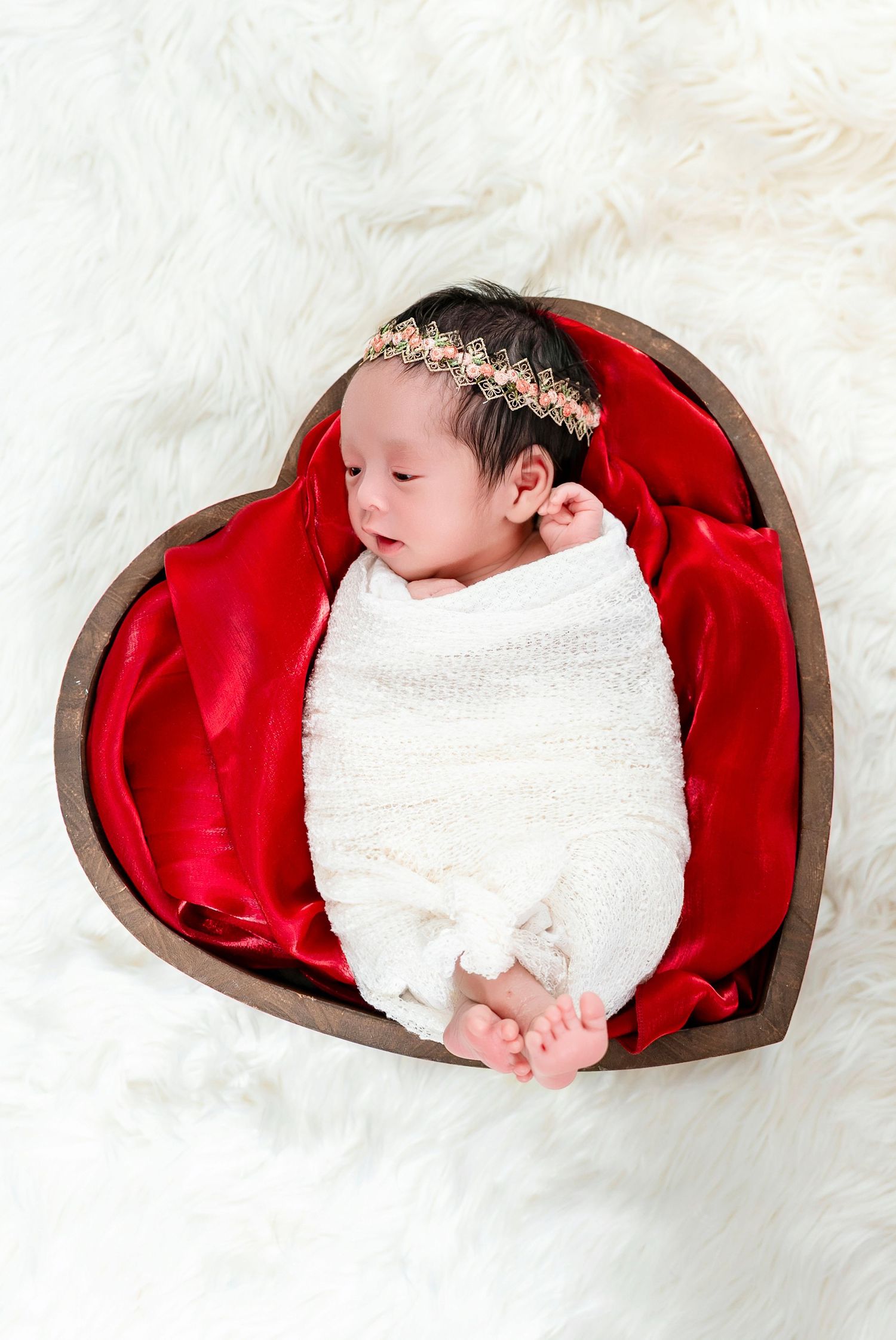 Sleeping newborn wrapped in white blanket inside heart-shaped basket with red satin lining.