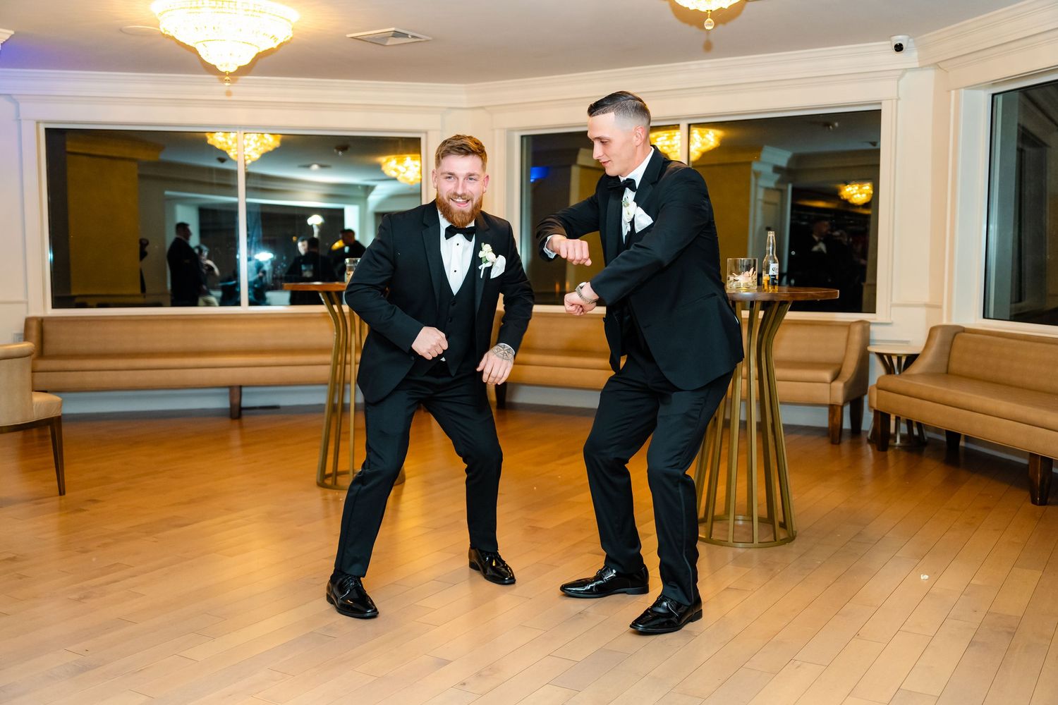 Groomsmen in black tuxedos perform choreographed dance moves on a wooden dance floor.