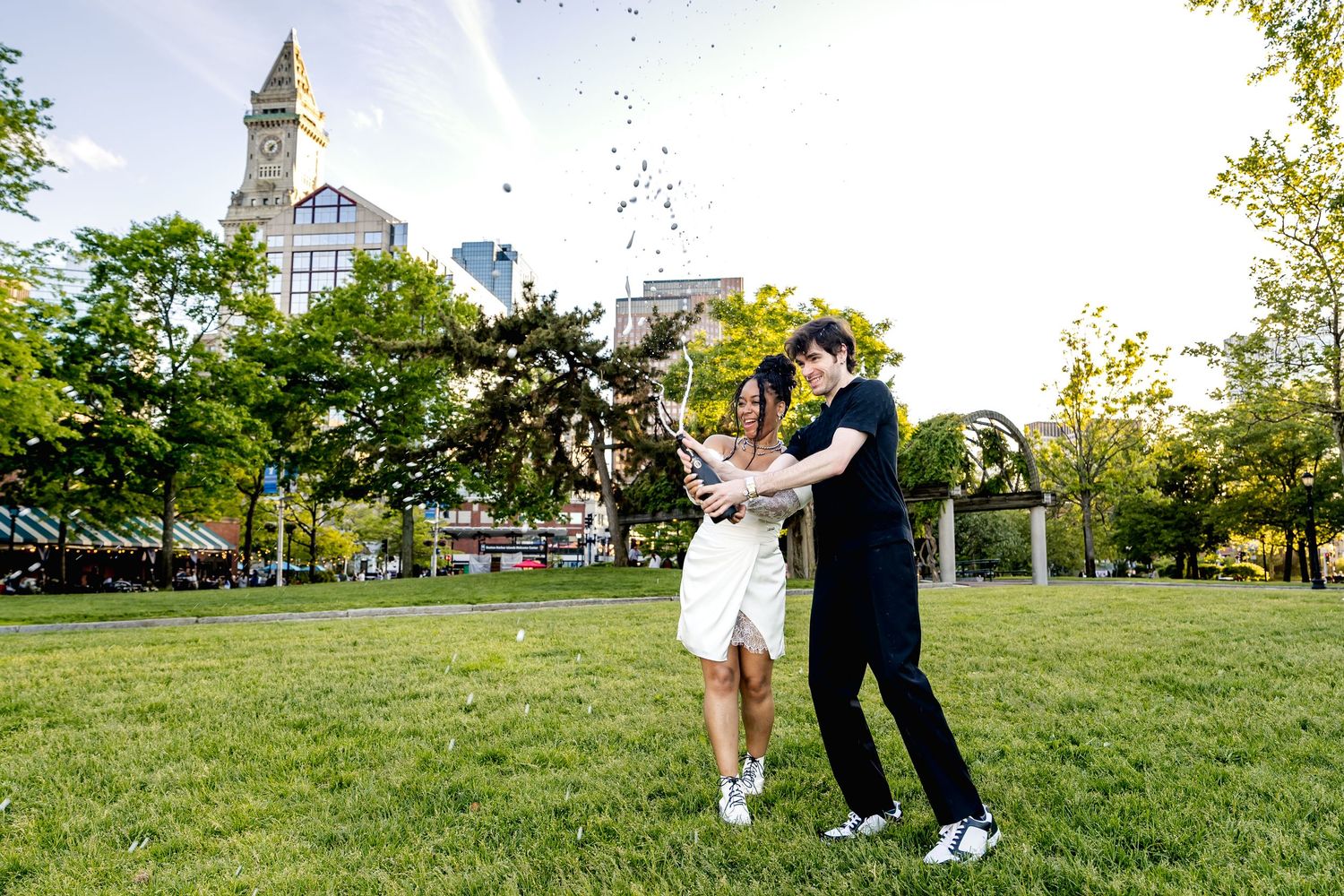 Couple joyfully tossing confetti in a green park with Boston Custom House clock tower in the background.