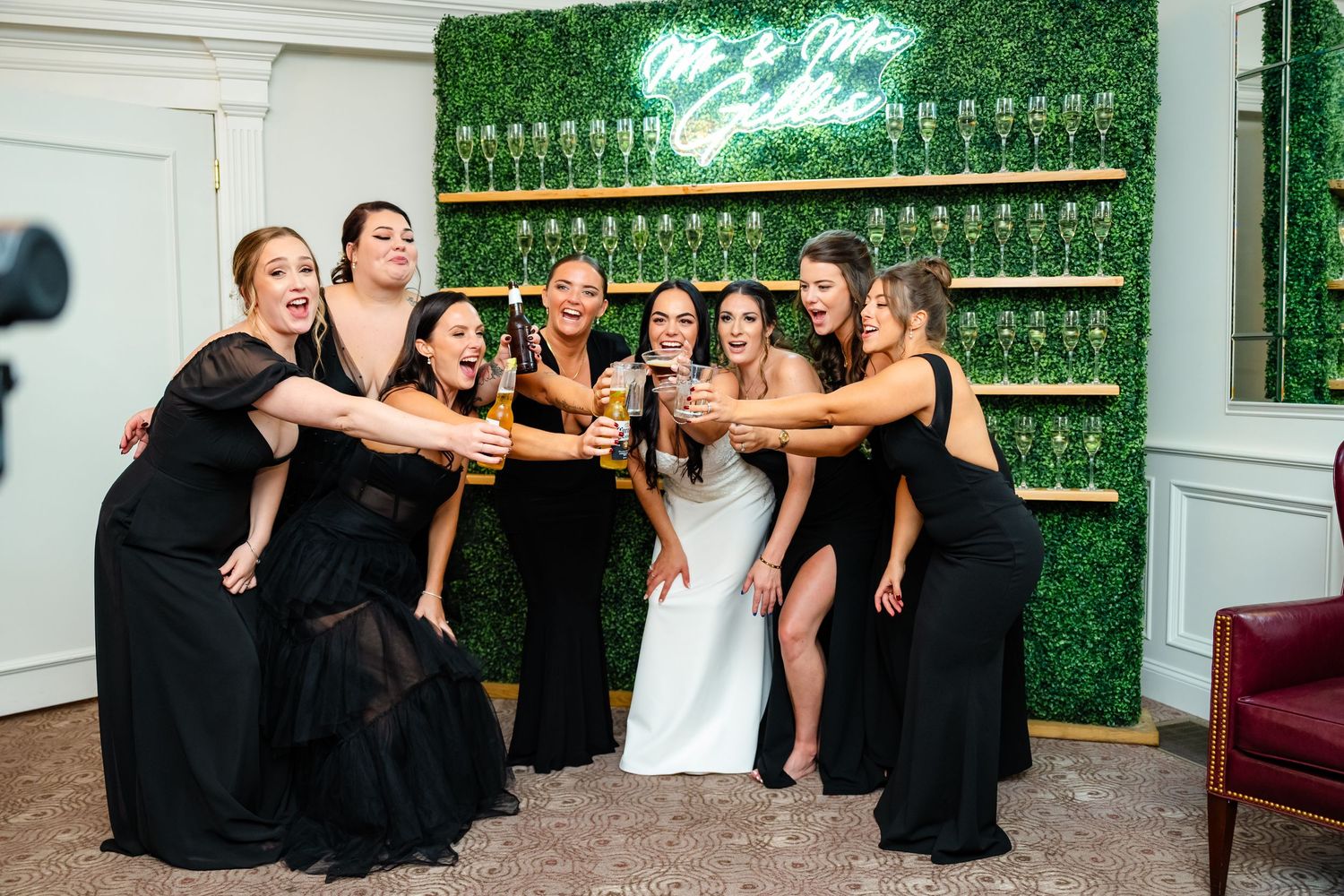 Wedding party celebrates with champagne toasts against a green hedge wall with glowing neon sign backdrop.
