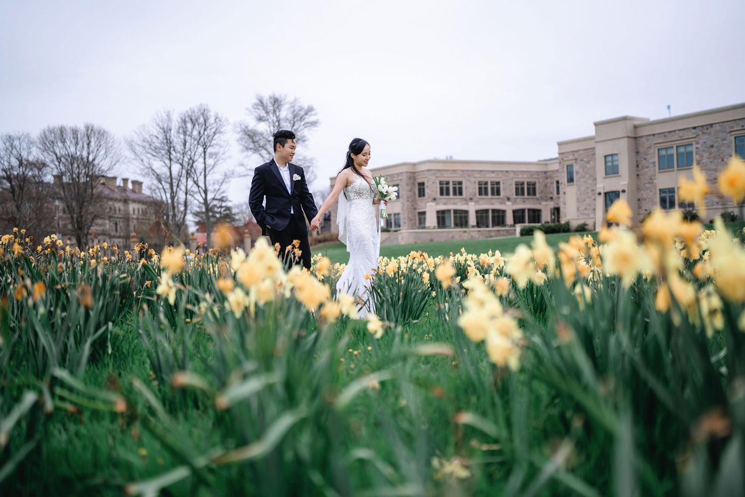 A couple walks through a spring garden with blooming yellow daffodils and historic buildings in the background.