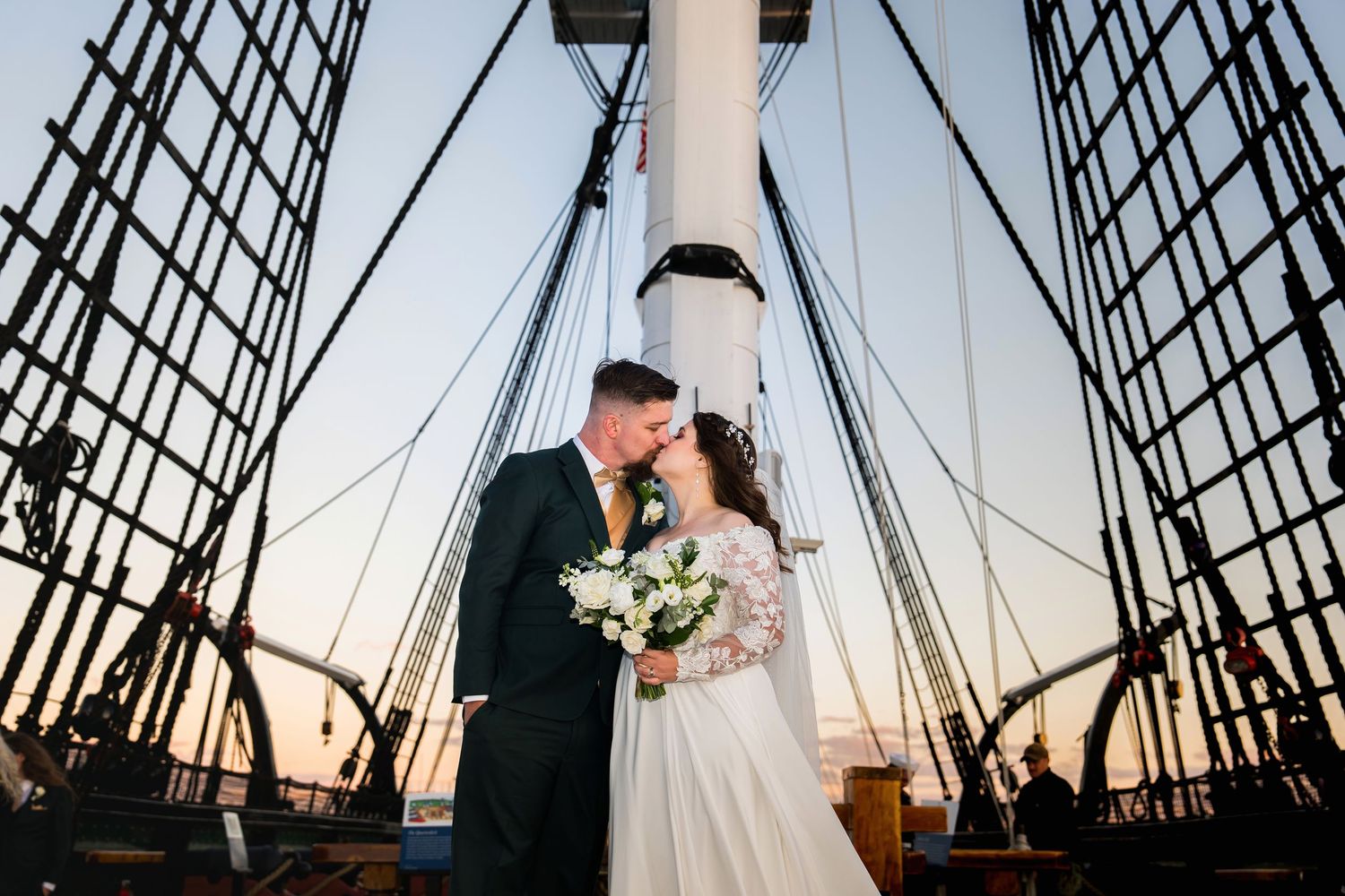 Newlywed couple embraces on a sailboat deck with rigging and masts silhouetted against a sunset sky.