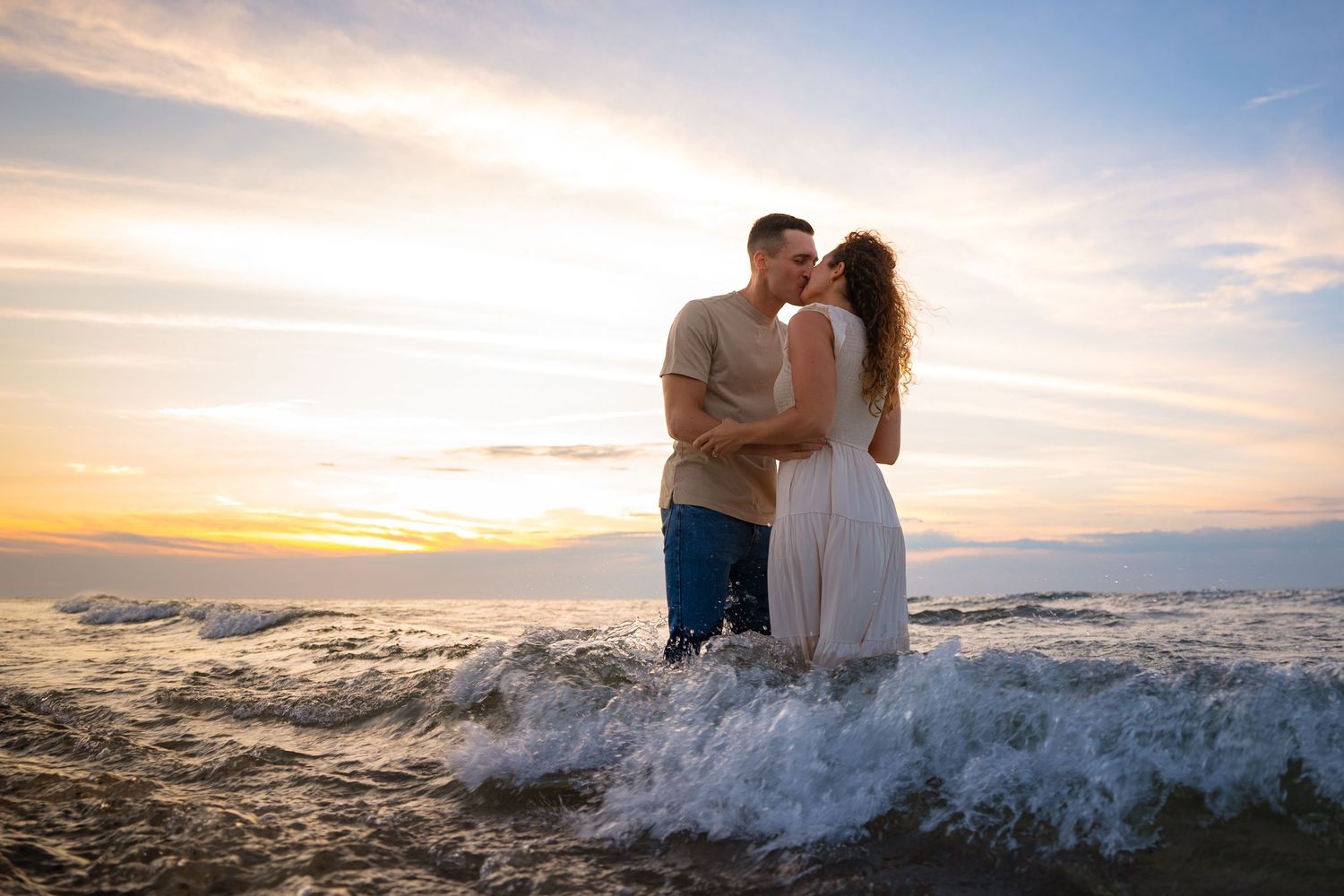 A romantic couple embraces on a rocky beach at sunset as ocean waves crash against the shore.