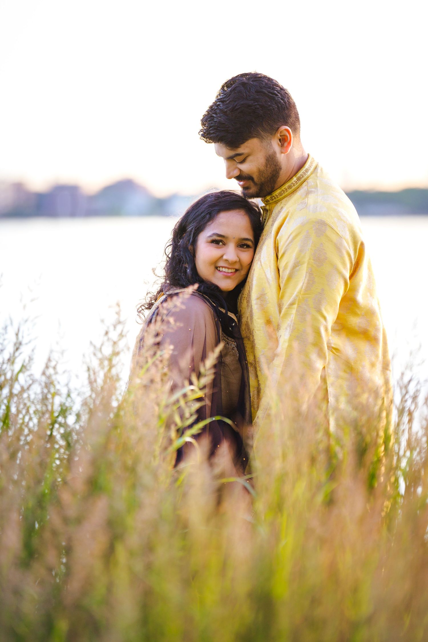 Romantic couple poses in tall grass during golden hour by the water.