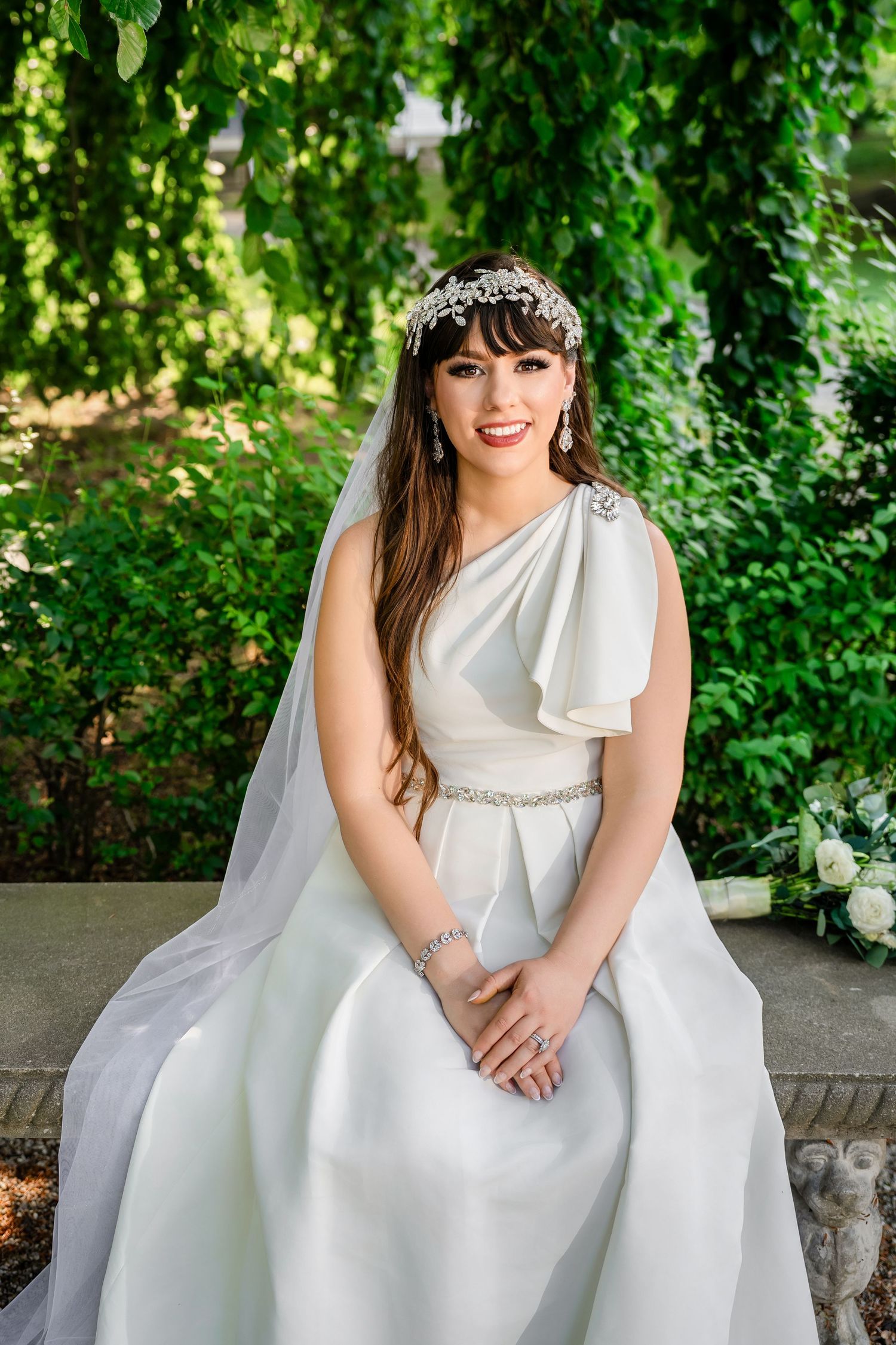 Bride wearing a classic Grecian-style white wedding dress with floral crown in garden setting.