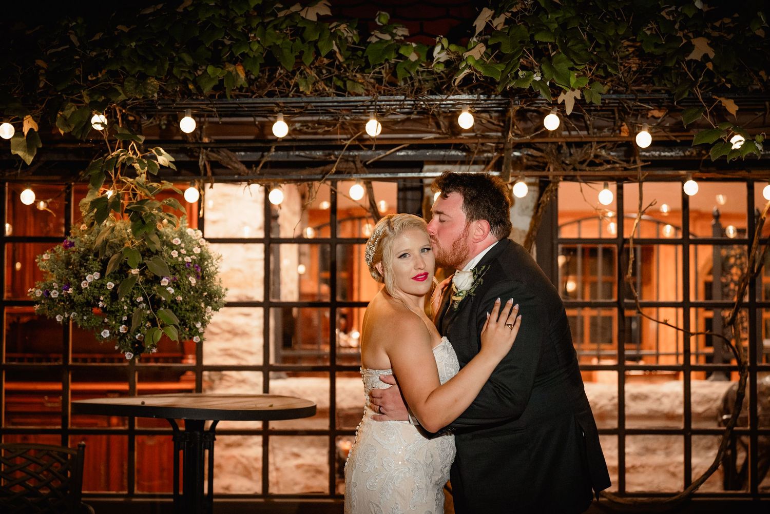 A romantic wedding couple shares a kiss under string lights at an outdoor evening venue.