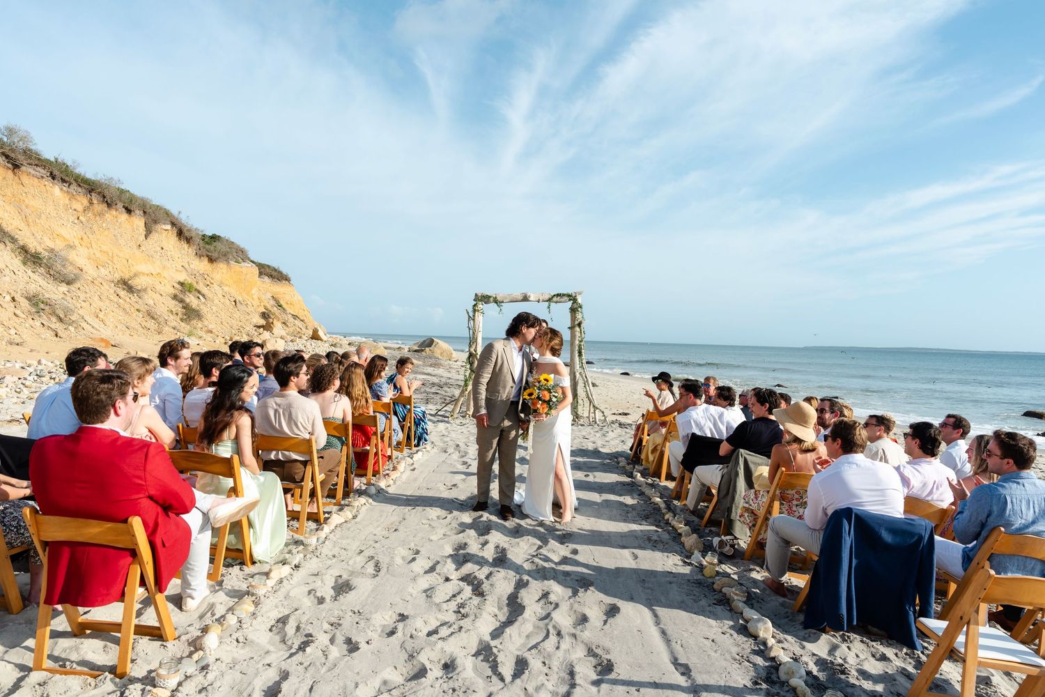 Panoramic view of a beach wedding ceremony with guests seated on the sand near coastal cliffs.