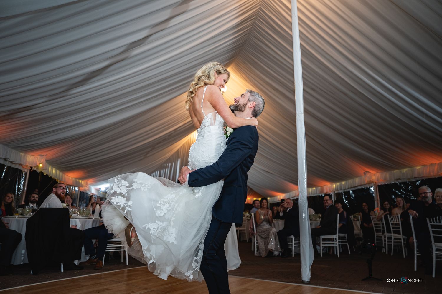 A couple shares their first dance under an elegant white tent at their wedding reception, with dramatic lighting and decor.