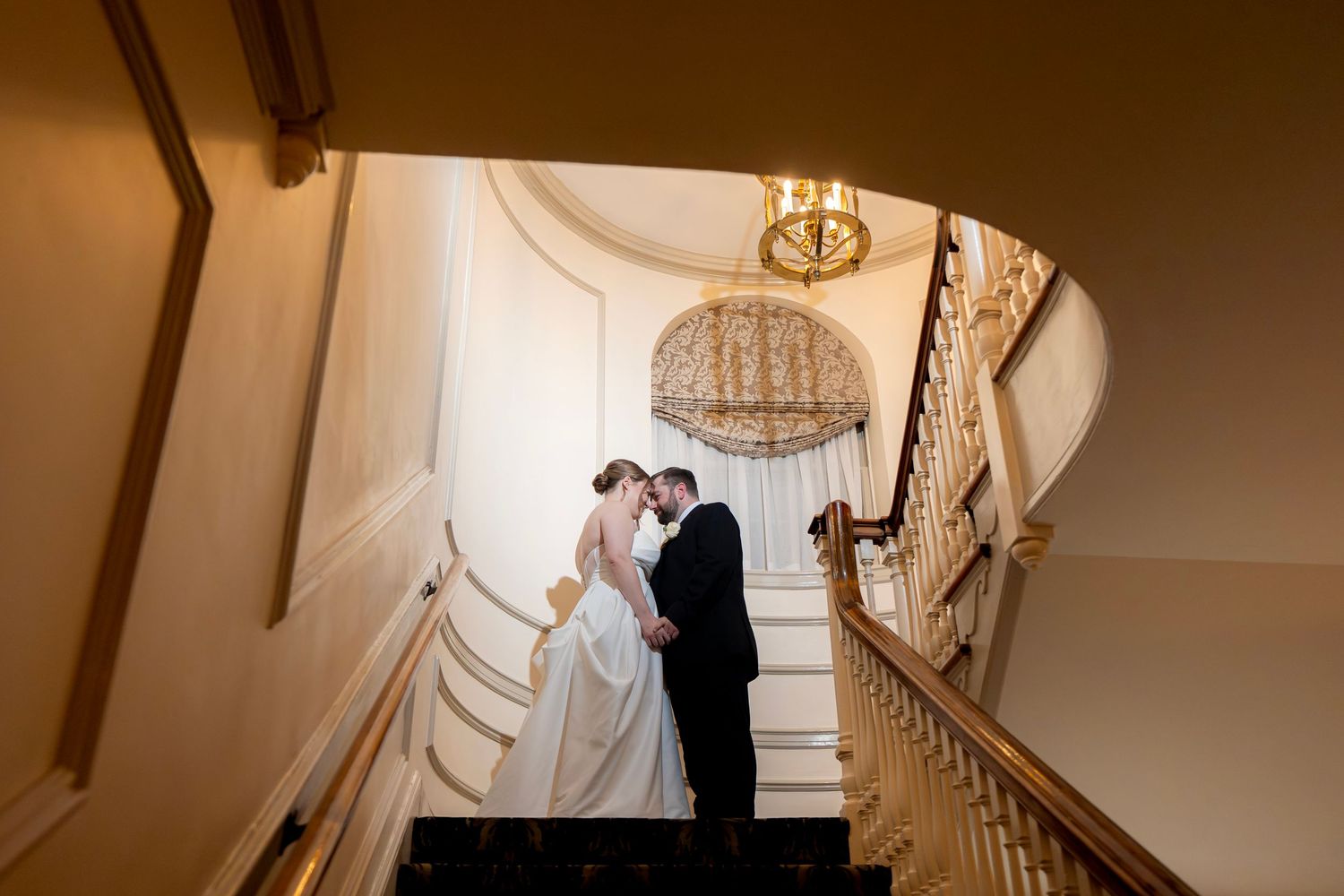 A wedding couple descends an elegant curved staircase in a grand venue with warm lighting and ornate details.