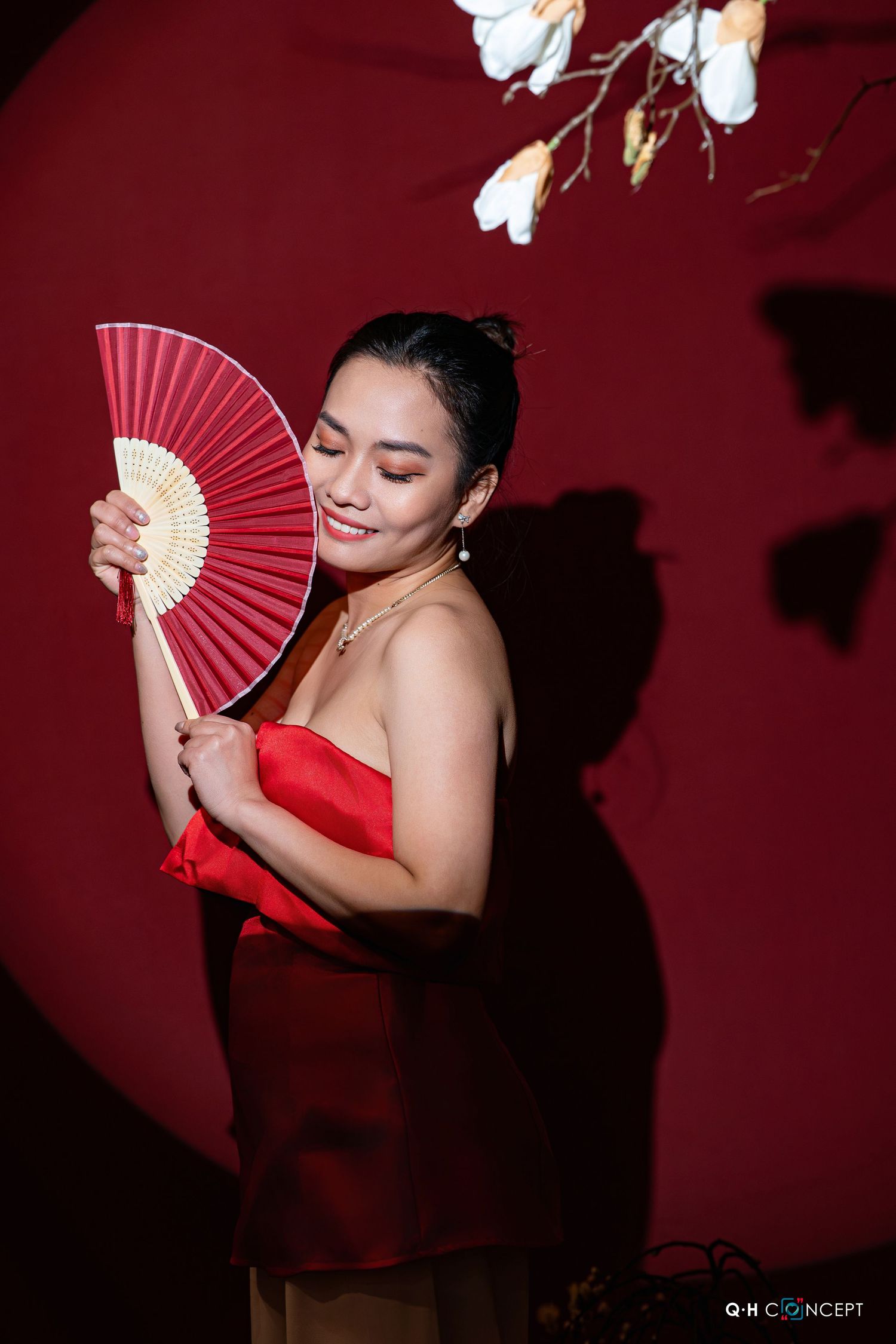 A figure in a red dress holds a decorative folding fan against a deep burgundy background with floral accents.
