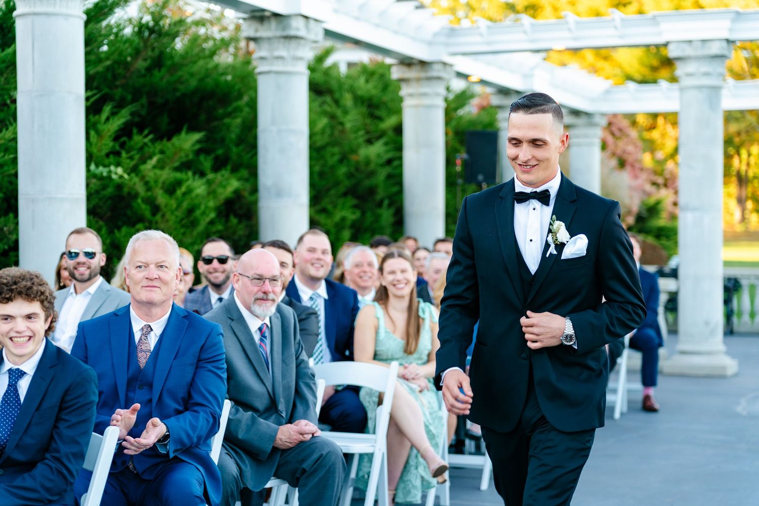 Wedding ceremony guests seated in white chairs under columned pergola during autumn.