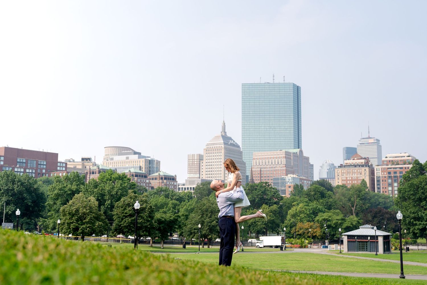 Couple embracing on green lawn in Boston Common with city skyline and skyscrapers in background.