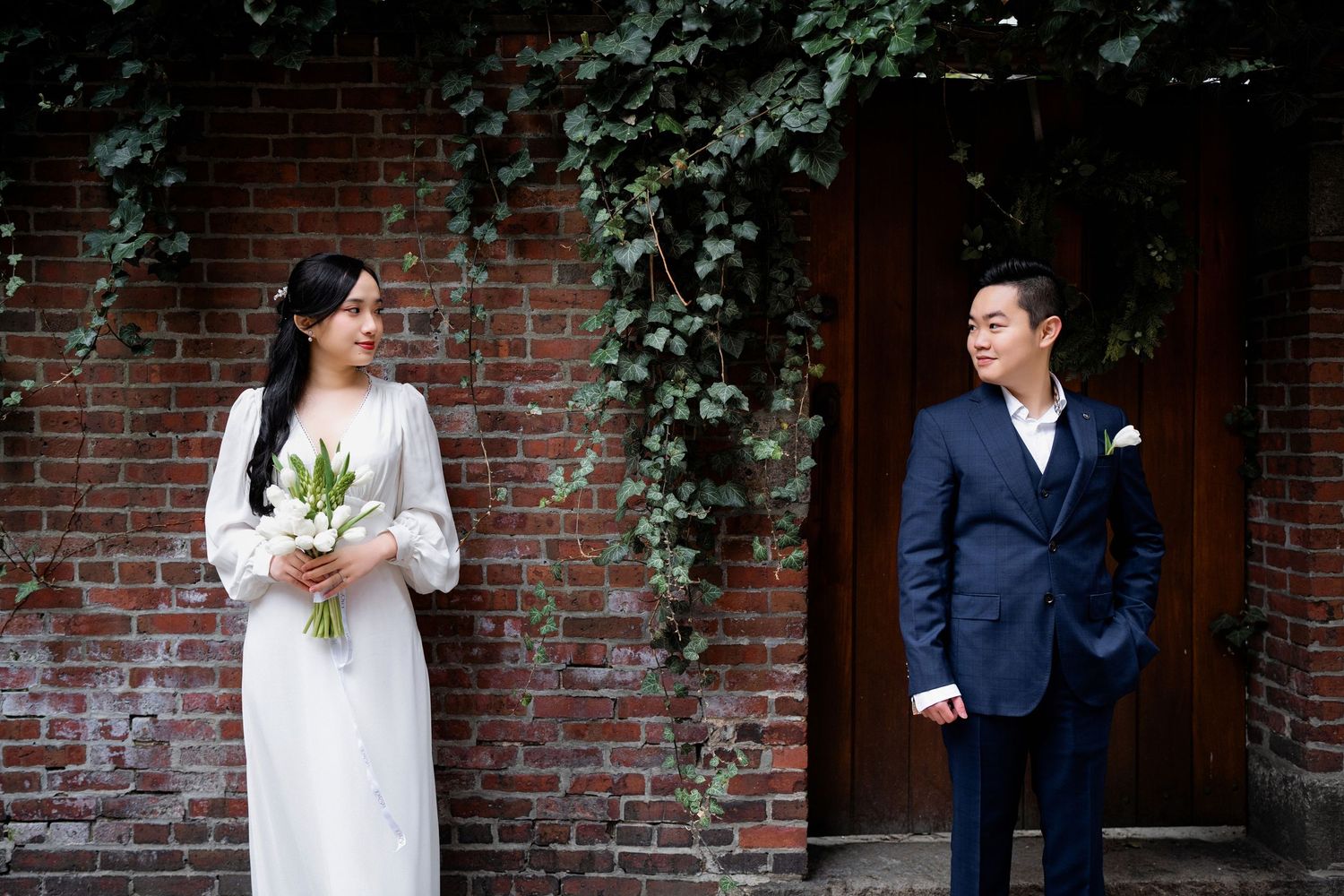 Wedding couple poses in front of rustic brick wall with greenery in romantic outdoor setting.