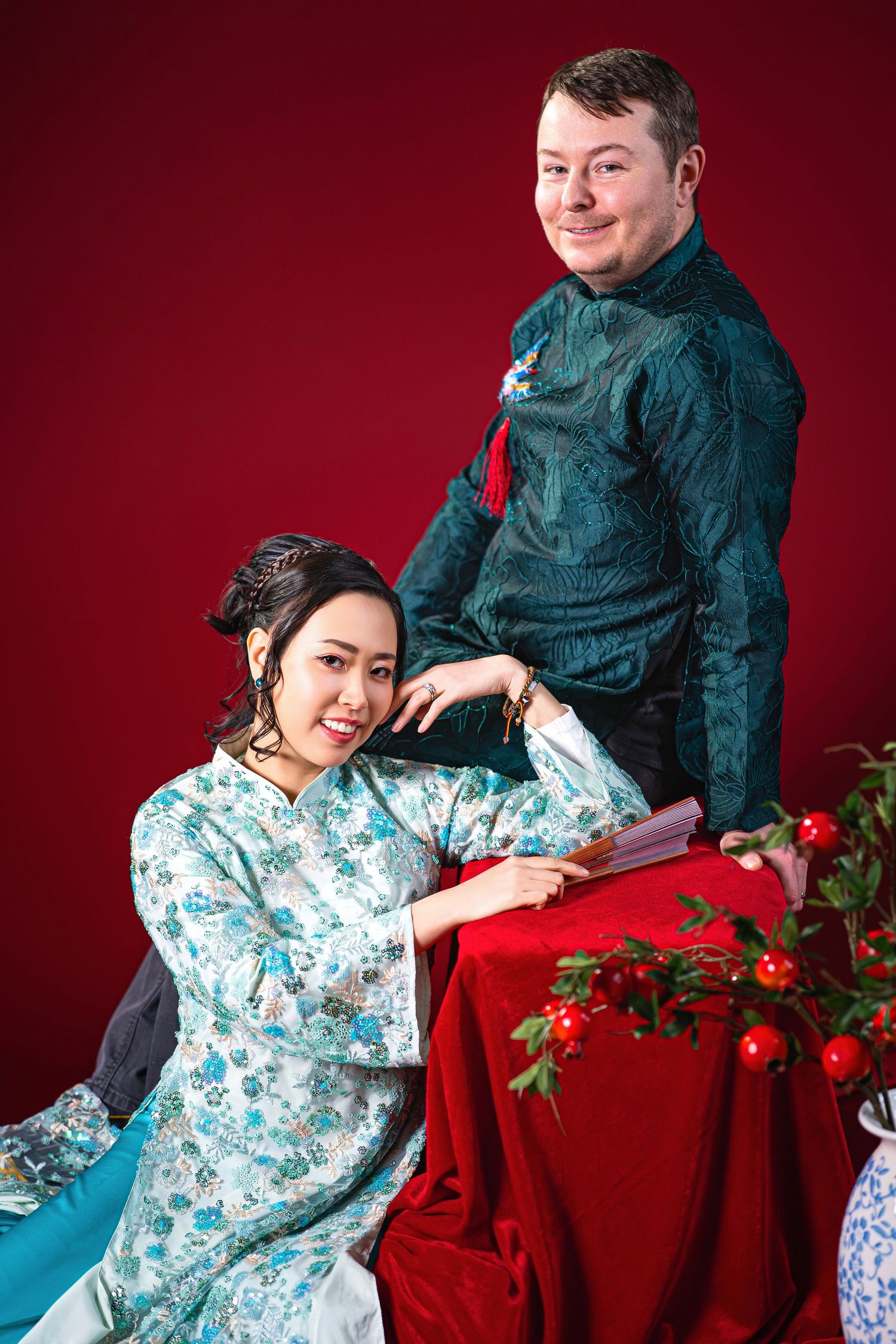 A couple poses together against a red backdrop with floral decorations in a romantic studio photoshoot.