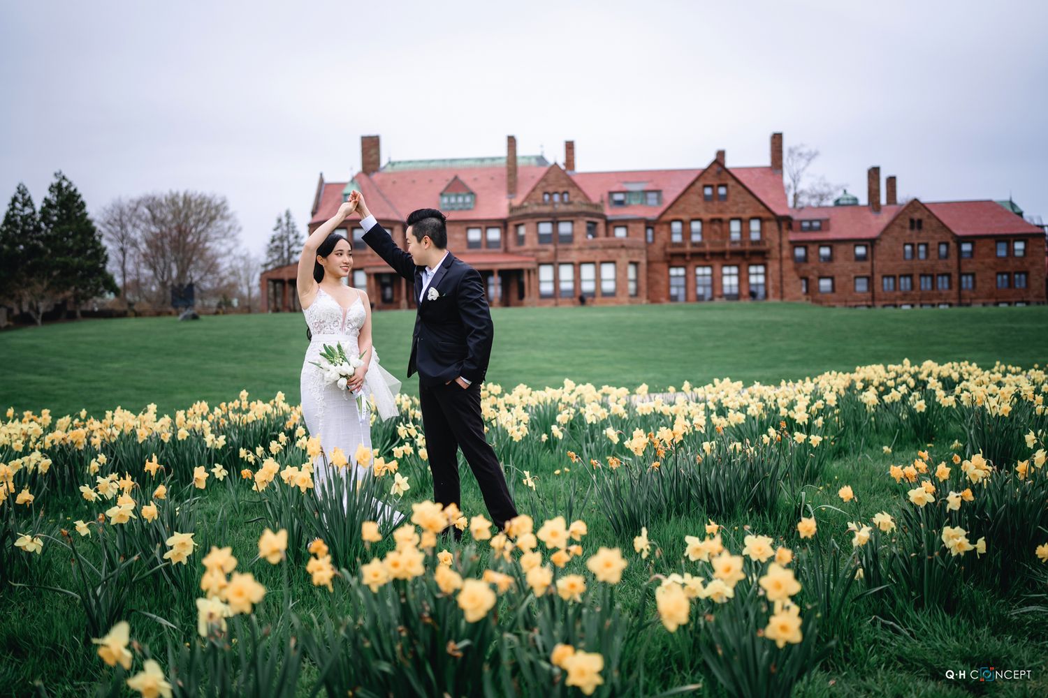 Couple poses for romantic engagement photos in a blooming spring daffodil field near historic brick buildings.